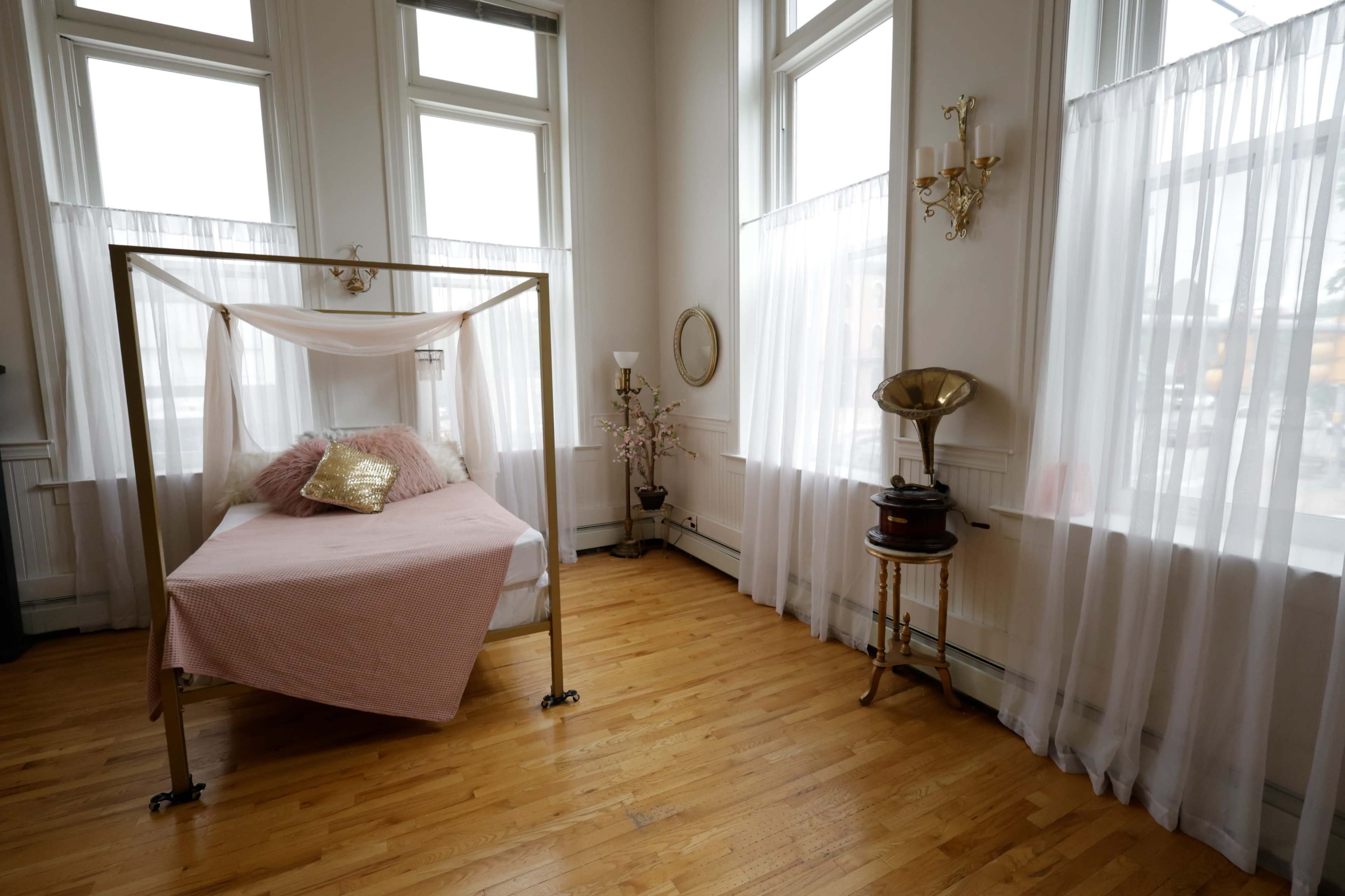 The room features a bed with a gold frame draped in sheer fabric, surrounded by large windows and elegant decor.