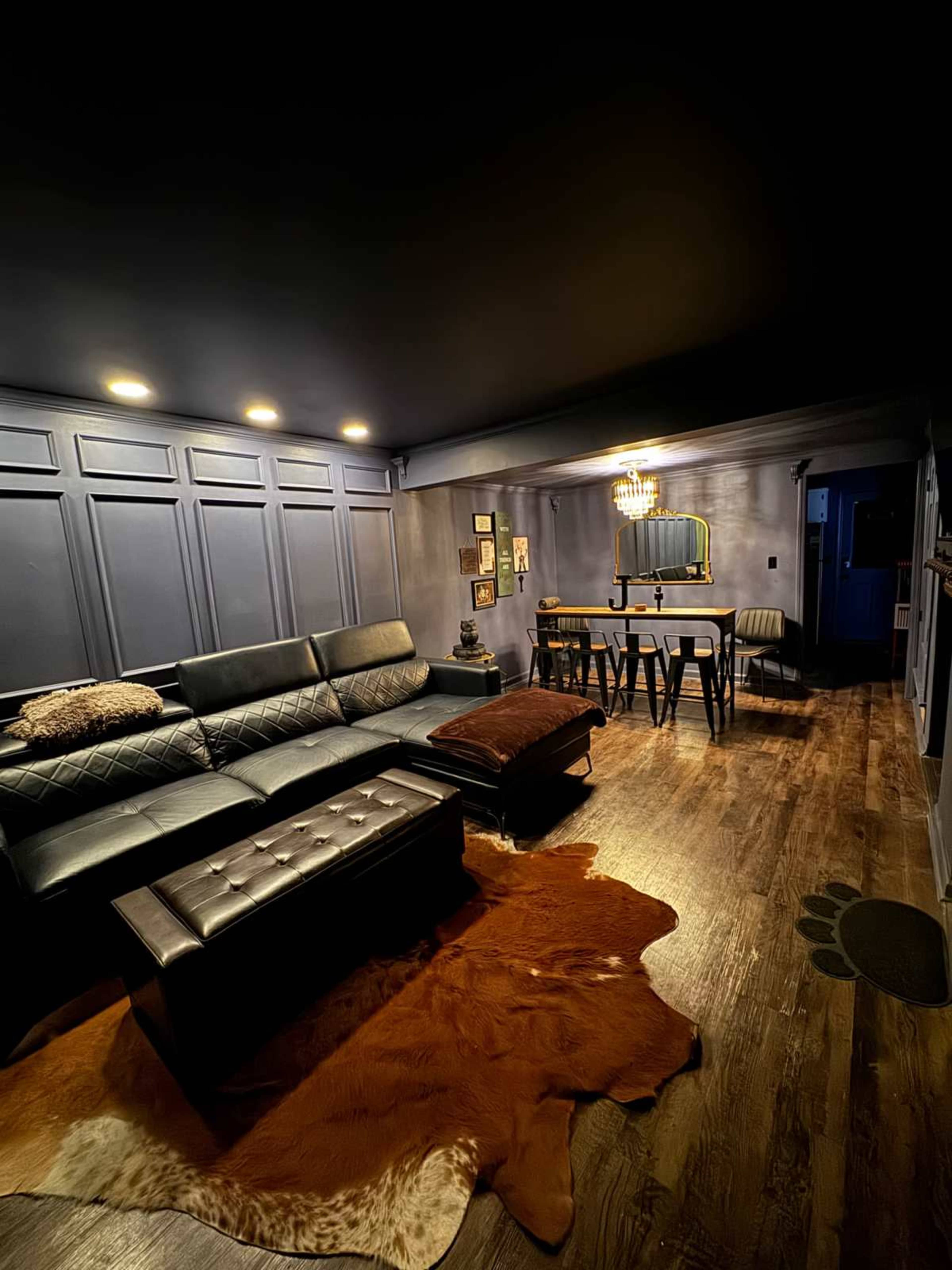 The image shows a darkly lit room featuring a black sectional sofa, a wooden dining table with stools, and a cowhide rug on the floor.