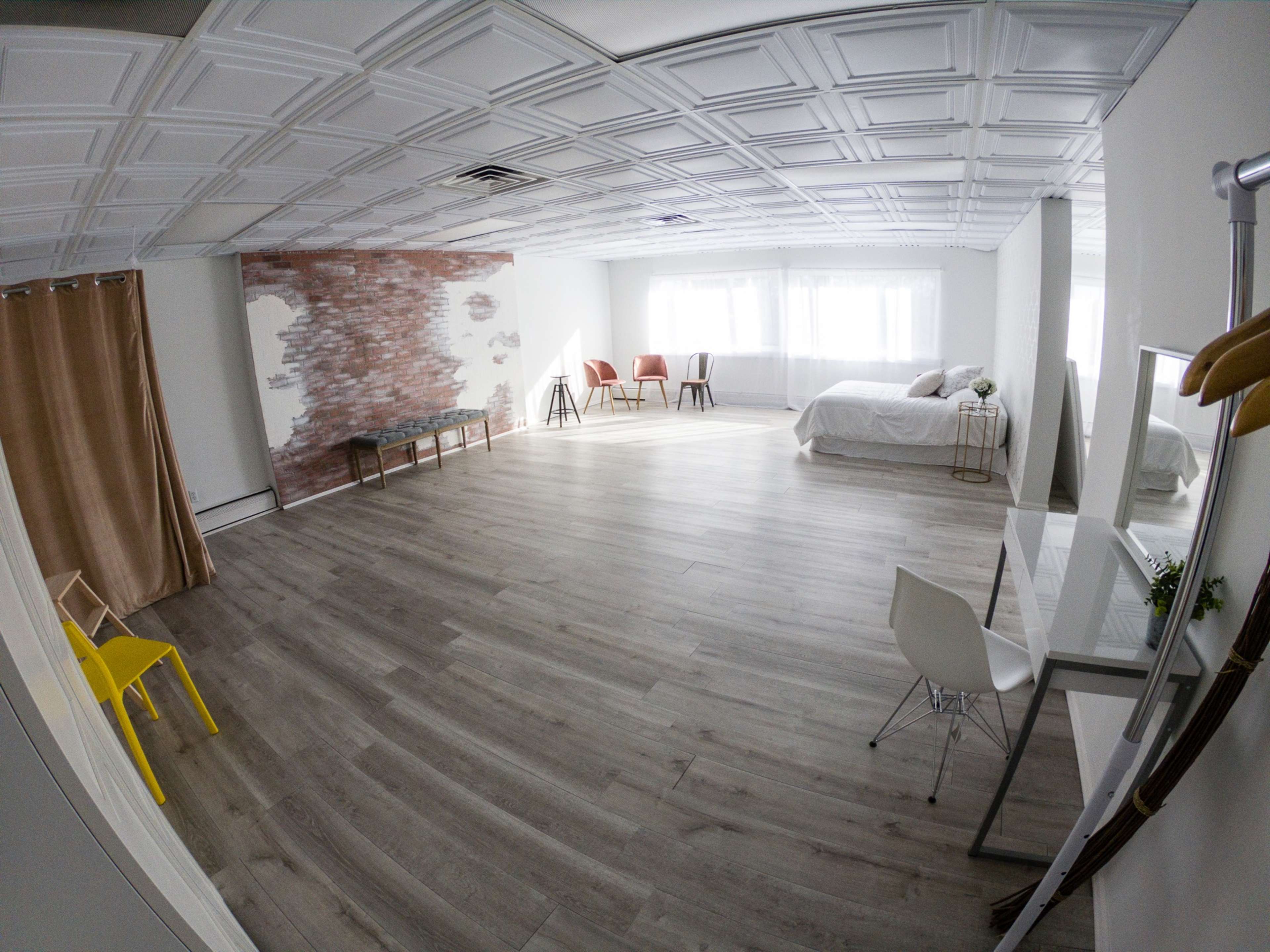 A spacious, well-lit room featuring a bedroom with a bed, a minimalist dressing area, and several chairs arranged around the space.