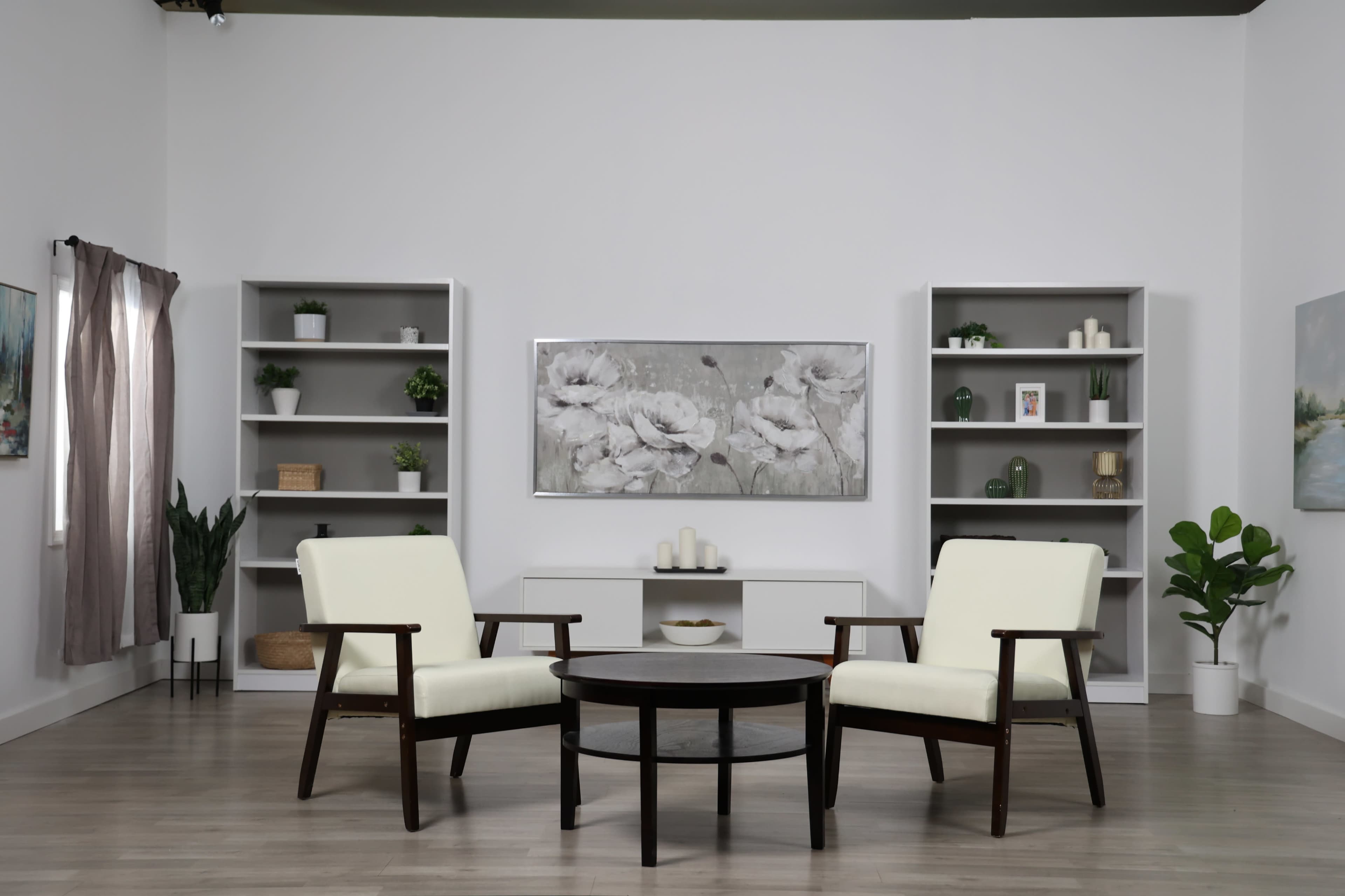 The image shows a minimalist living room setup with two cream-colored chairs facing a round table, surrounded by shelves displaying decor and plants.