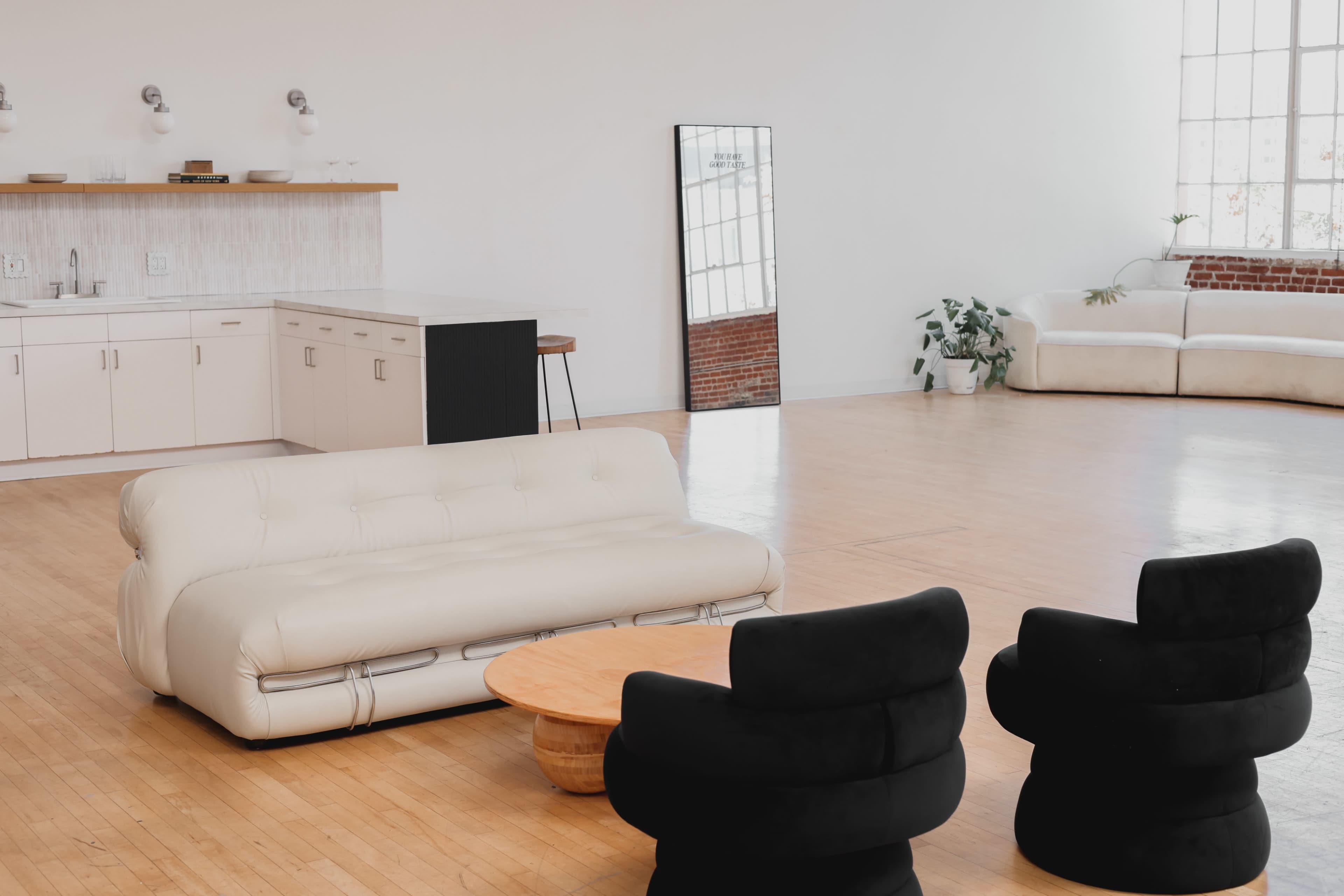 The image shows a spacious room with a white sofa, two black chairs, a round wooden table, a kitchen area, and large windows allowing natural light.