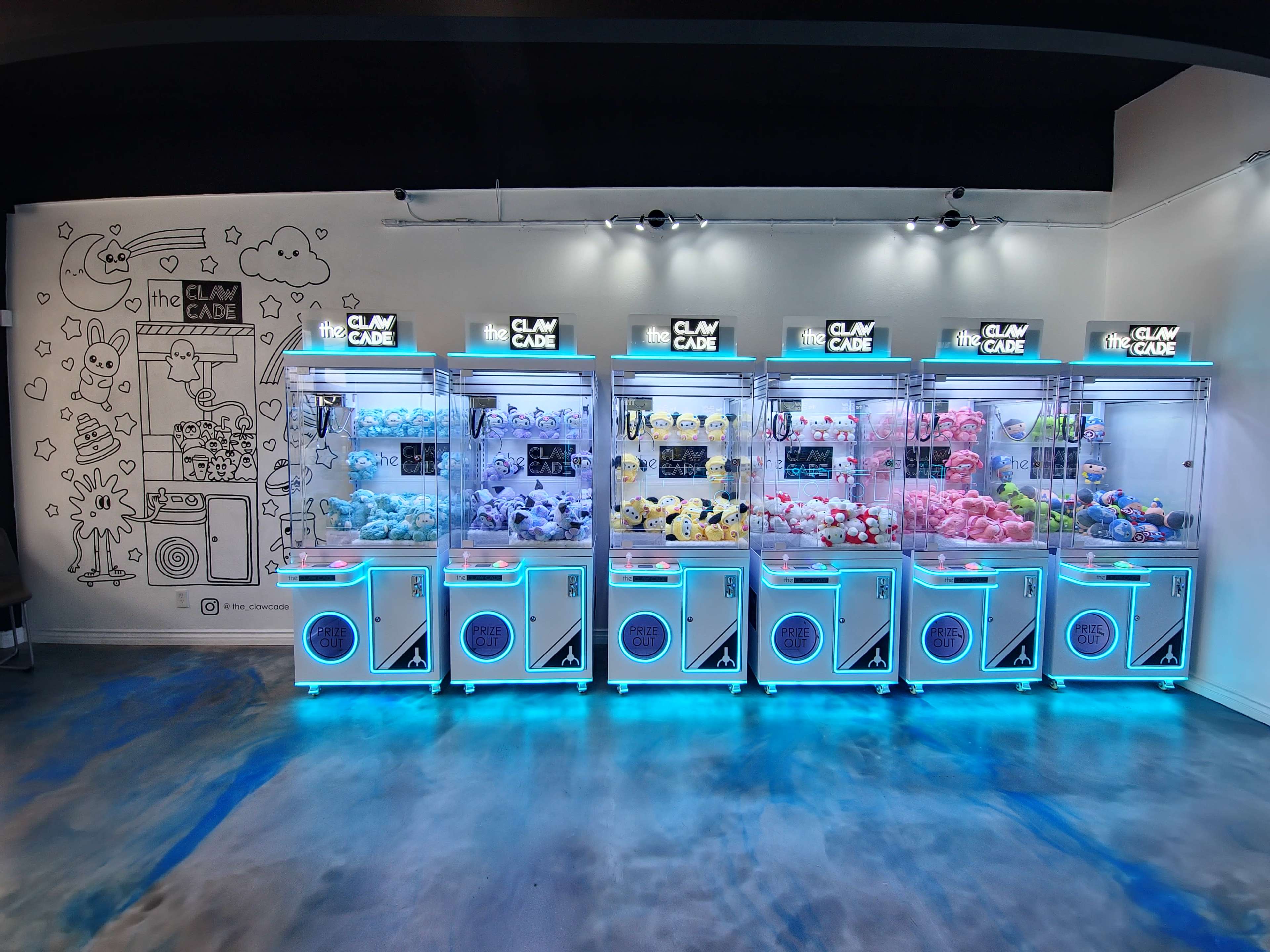 The image shows a row of brightly lit claw machines filled with various plush toys against a decorated wall with cartoon illustrations.