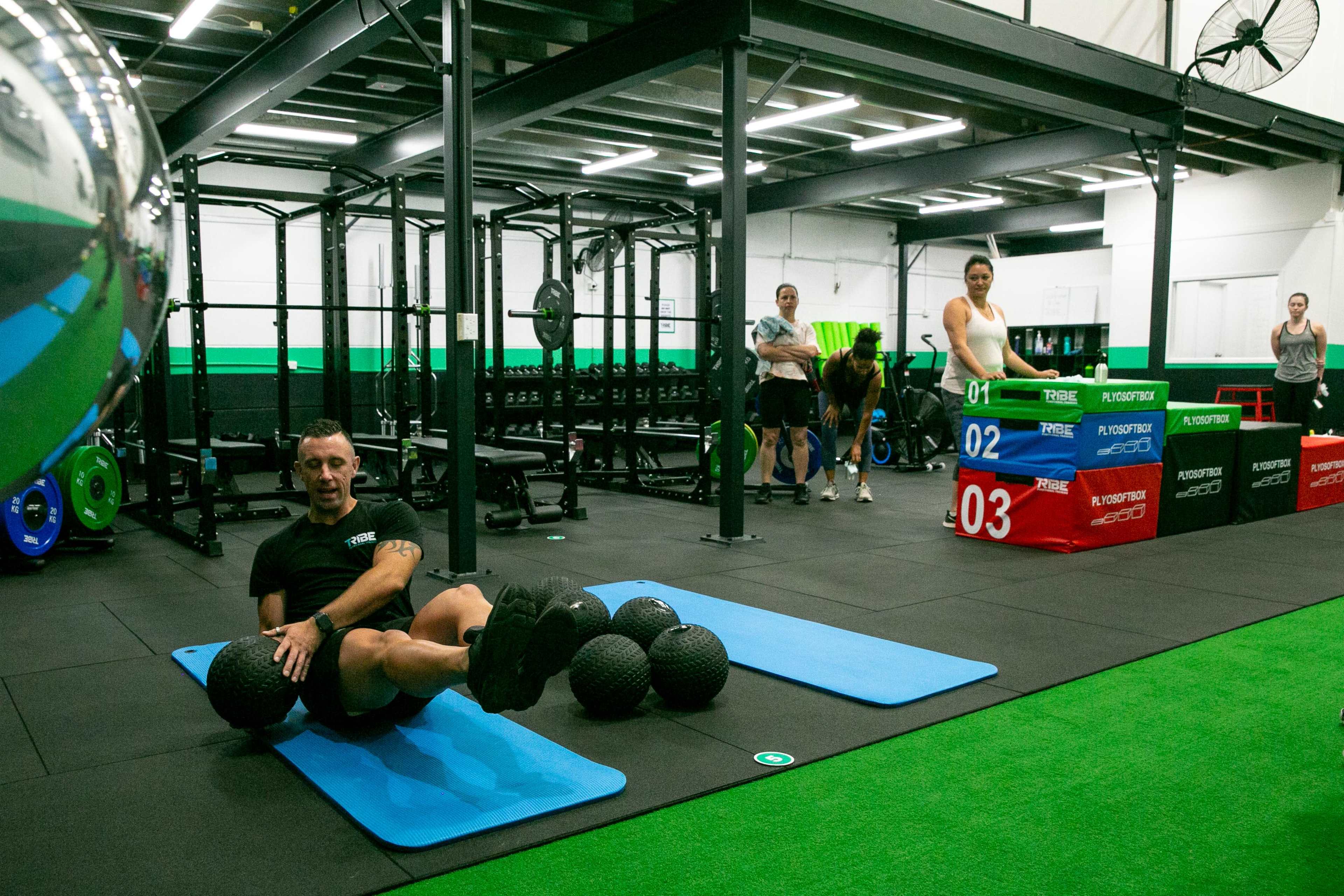A fitness studio features individuals engaged in various exercises, with one person performing seated ball exercises on a mat in the foreground.