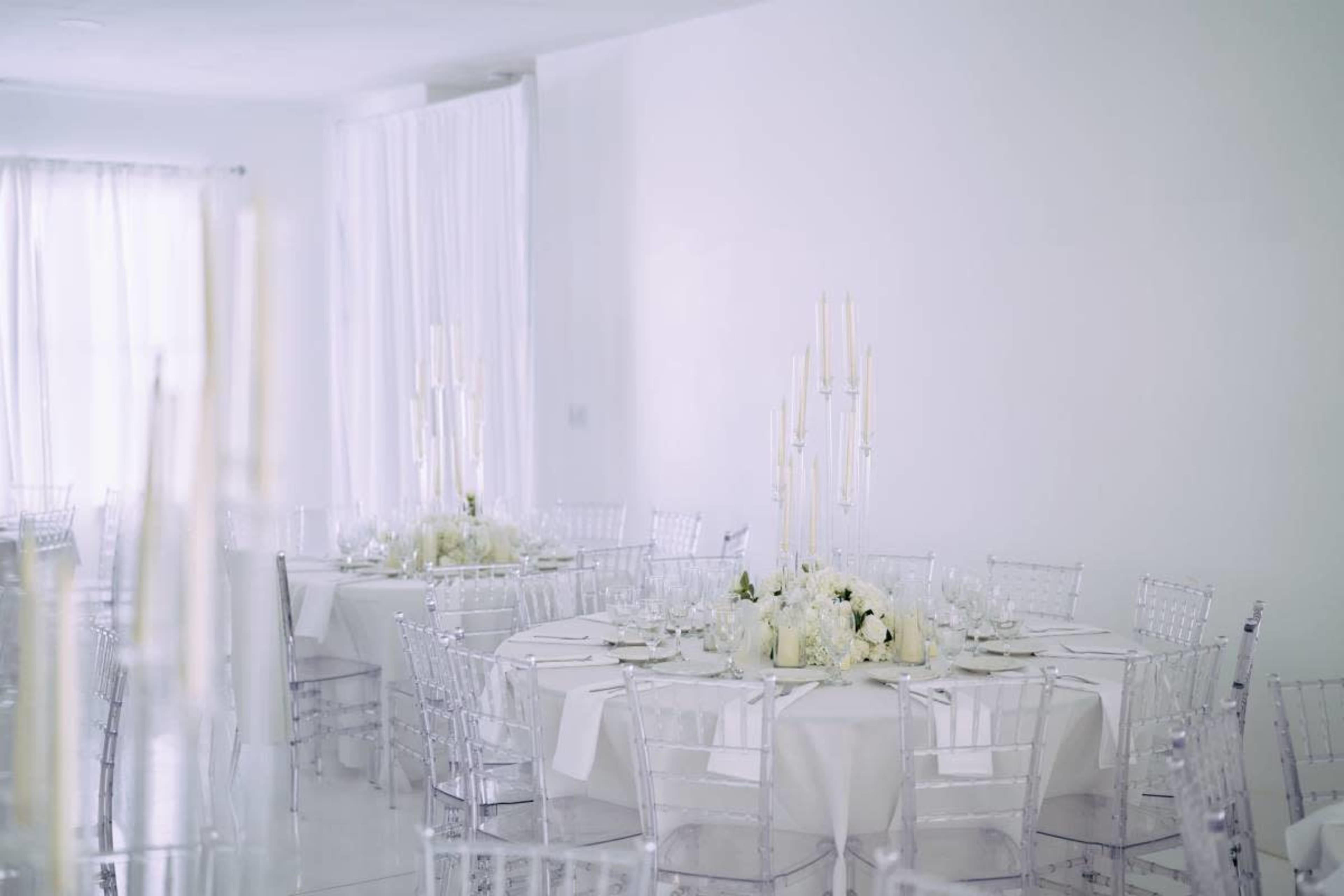 A bright, white banquet hall features circular tables adorned with floral centerpieces and tall candlesticks.