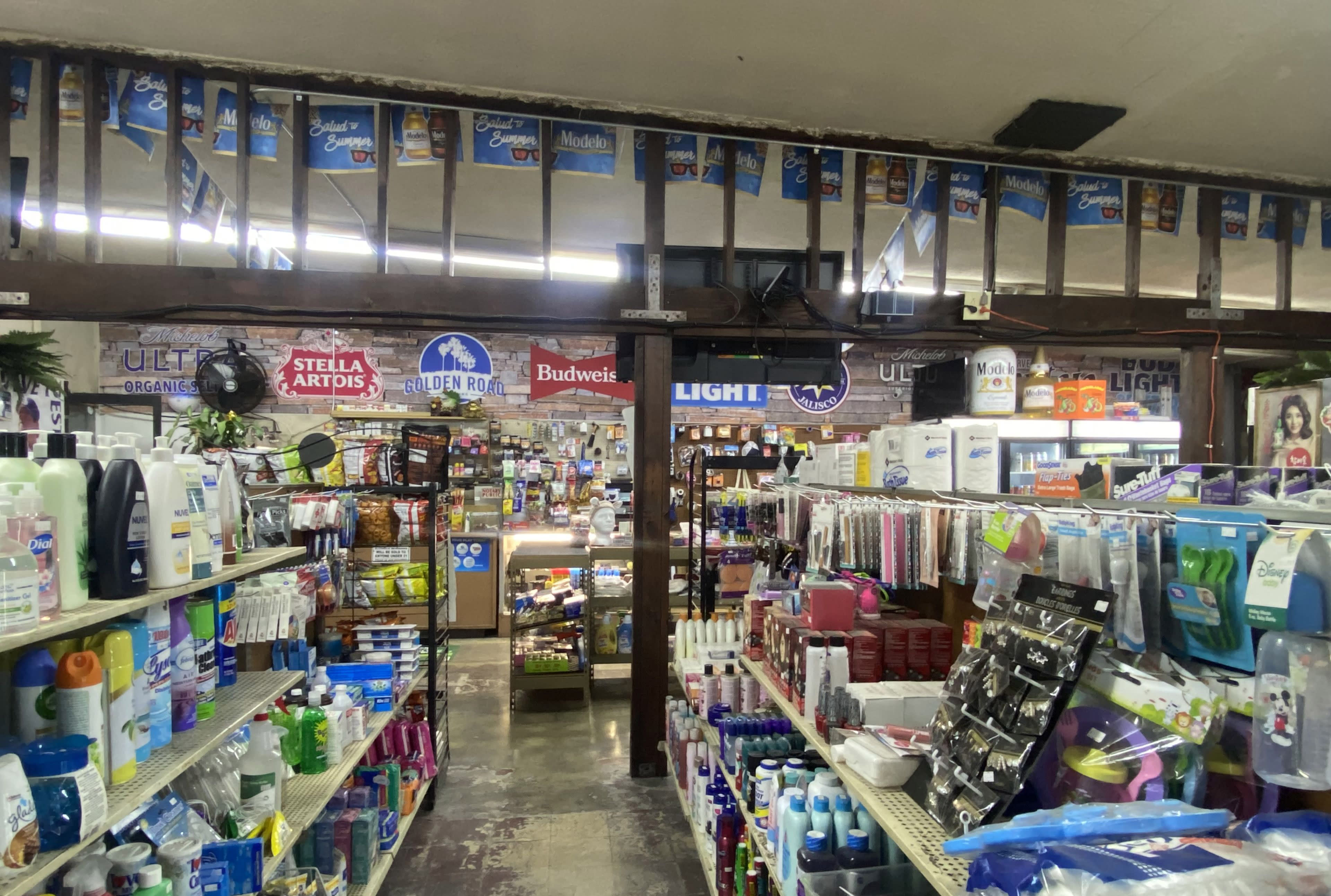 The interior of a convenience store, featuring shelves stocked with various household products and signage for beer brands hanging above.