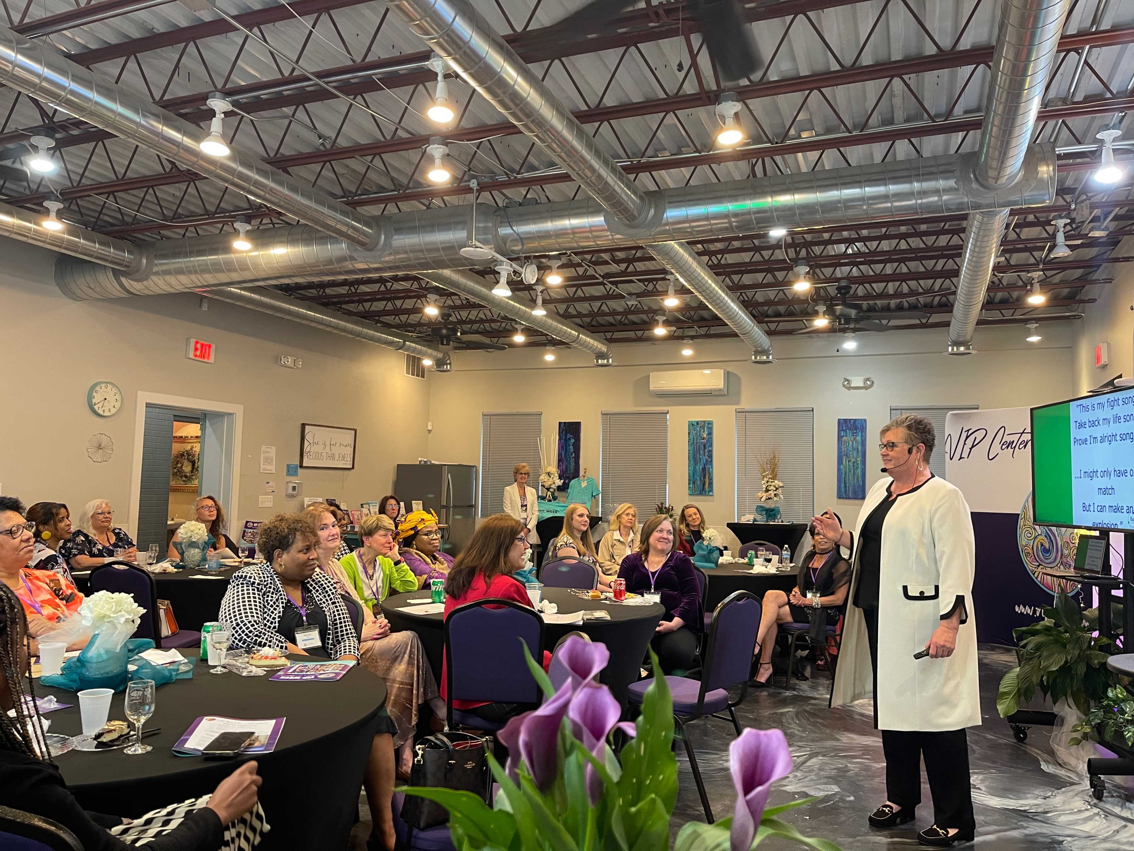 A speaker presents to an audience seated at round tables in a well-lit event space.