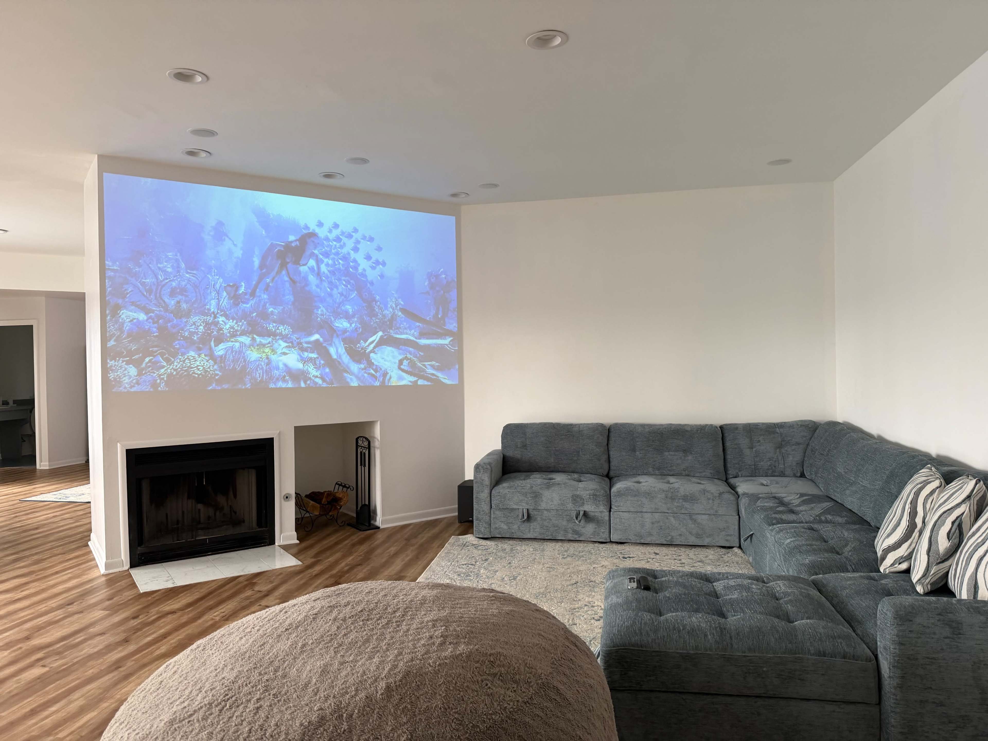 A projector displays an underwater scene on the wall of a modern living room with a large sectional sofa and a fireplace.
