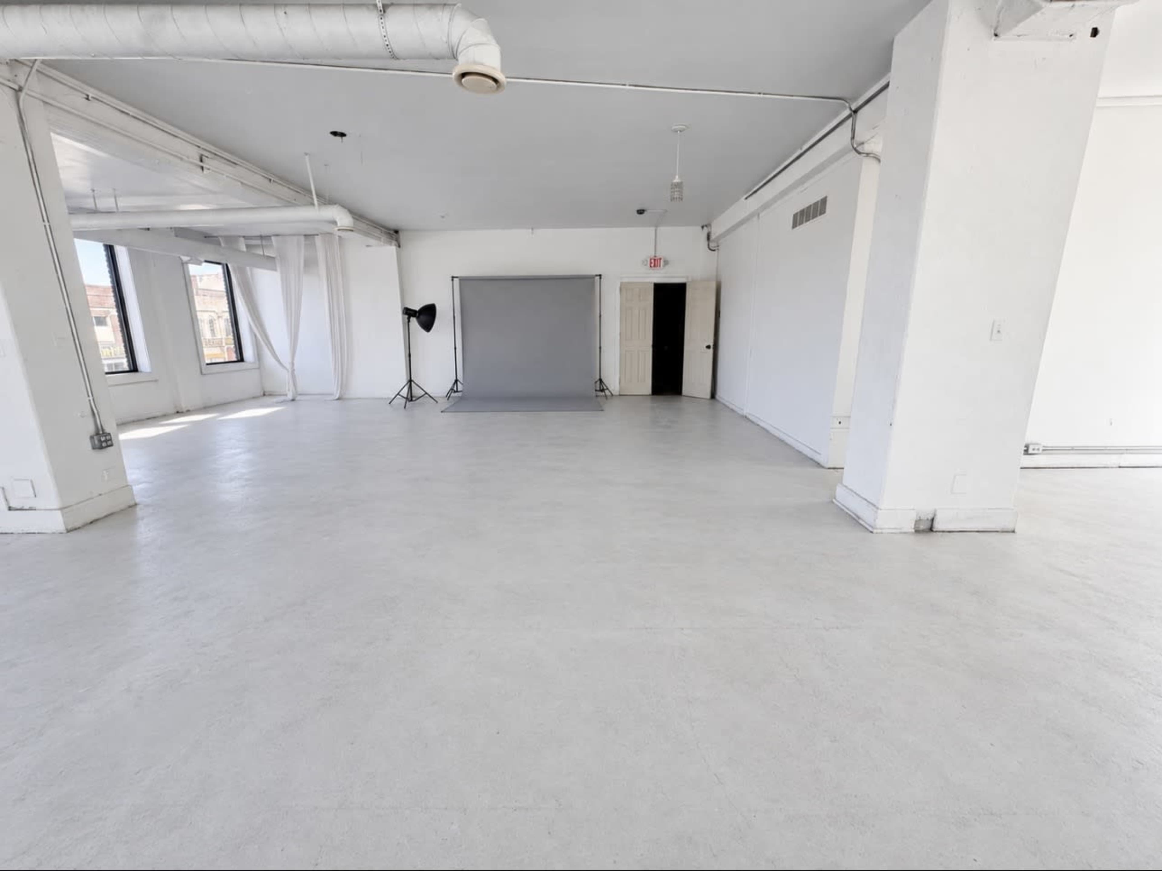 The image shows a spacious, empty room with white walls, a concrete floor, and large windows providing ample natural light.