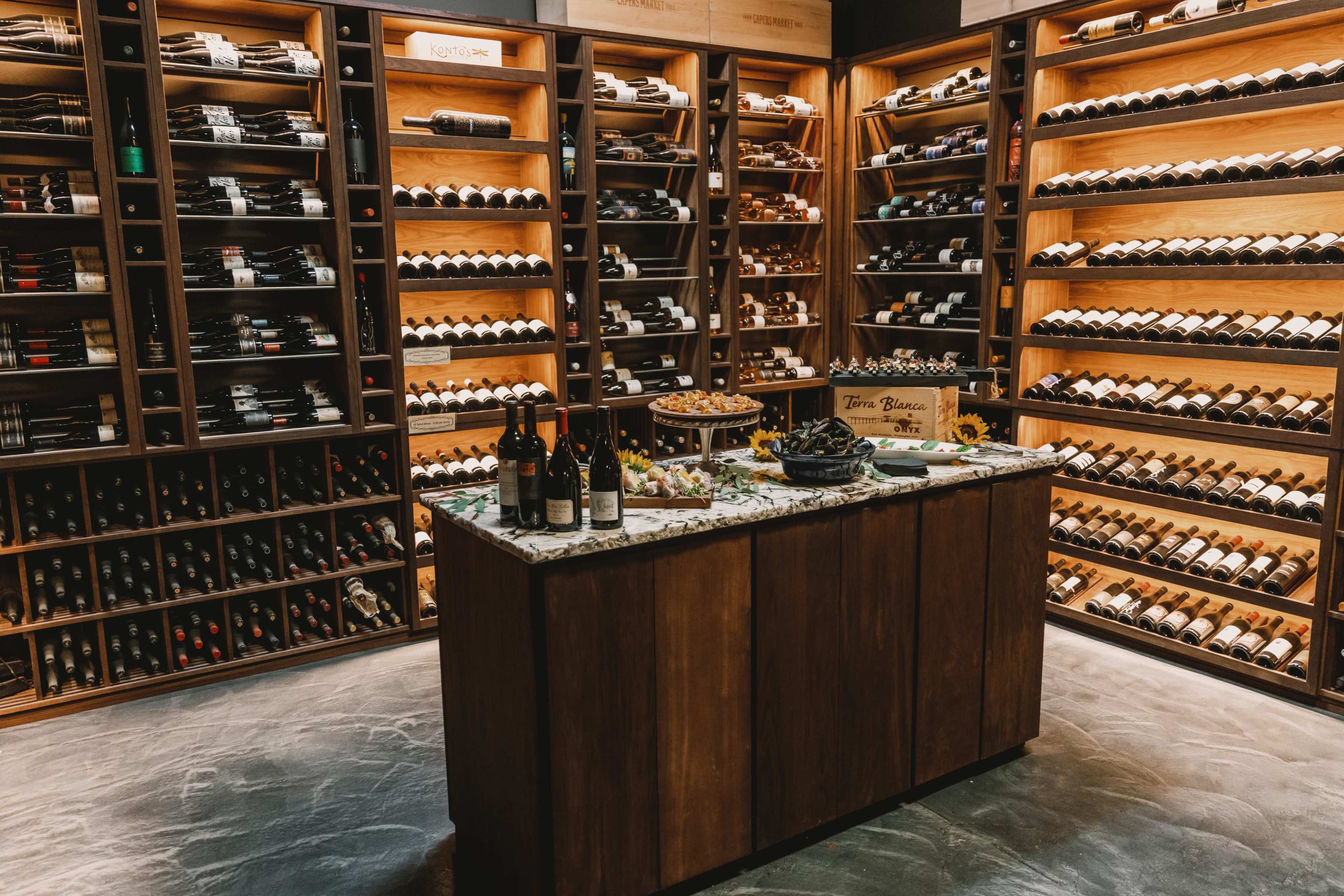 A wine cellar features wooden shelving filled with bottles of wine, with a marble-topped table in the center adorned with snacks and wine bottles.
