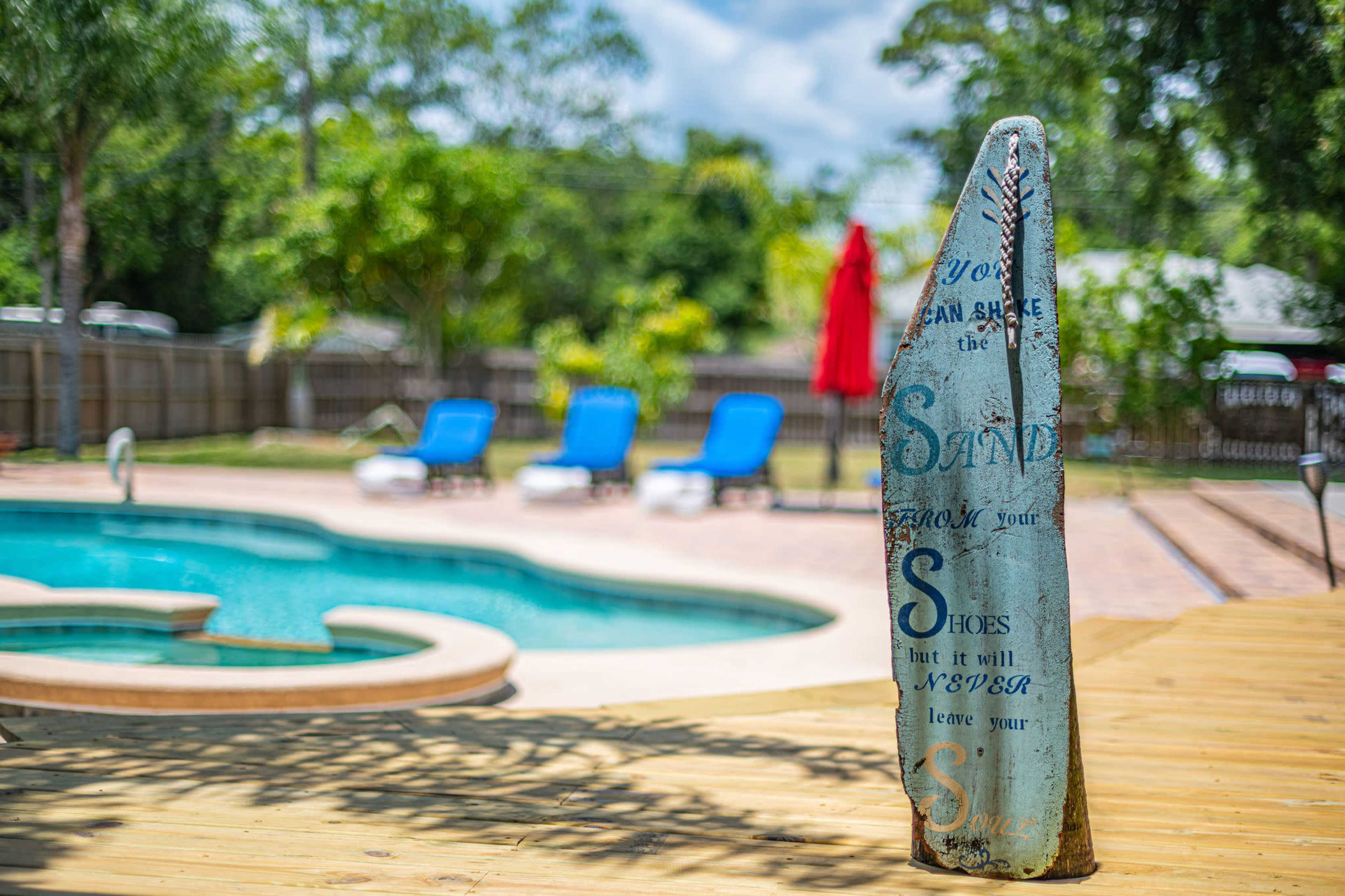 A decorative wooden sign stands next to a swimming pool with lounge chairs and an umbrella in the background.