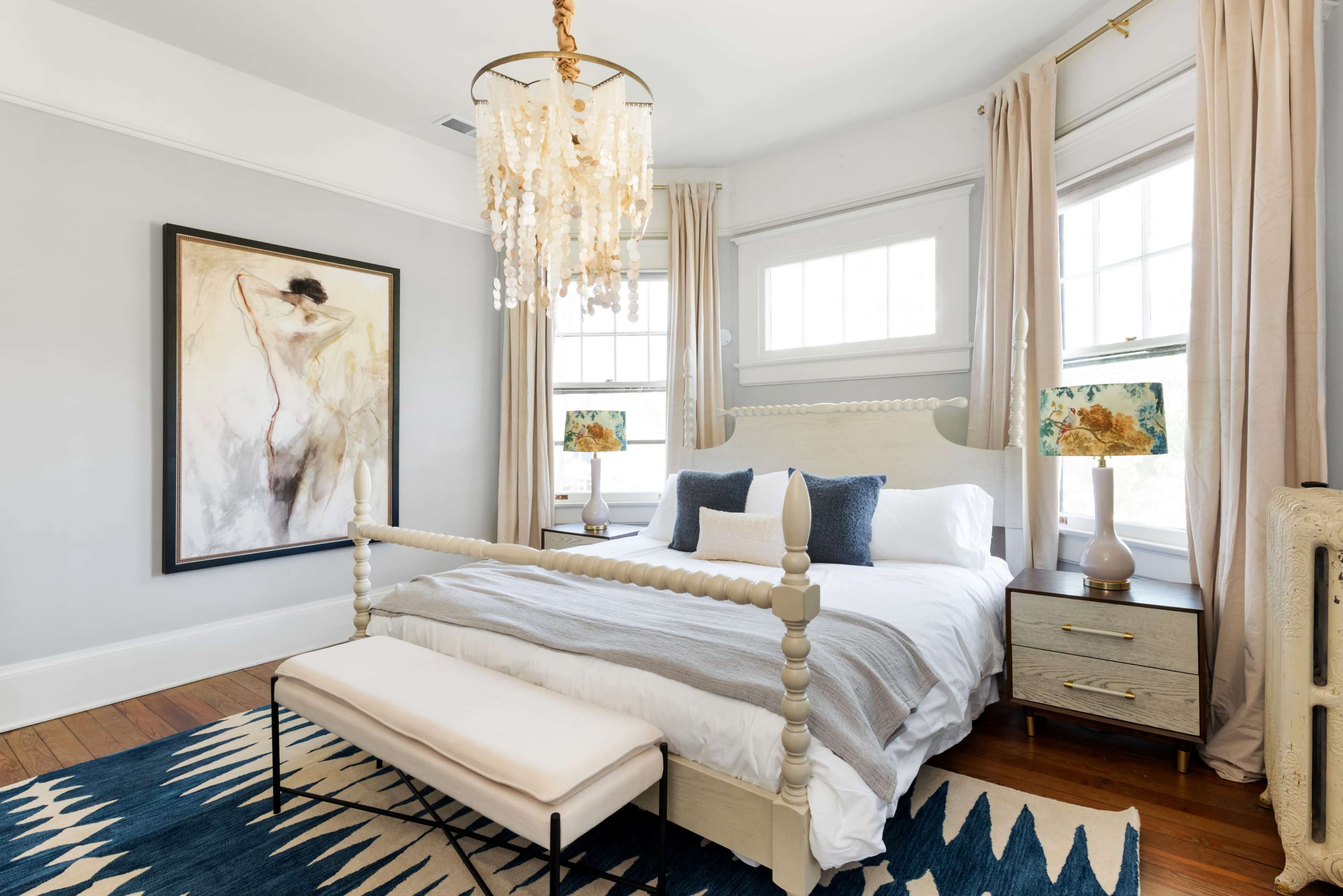 A well-decorated bedroom features a large bed with a light-colored headboard, a bench at the foot, and artwork on the wall, illuminated by a decorative chandelier and table lamps.