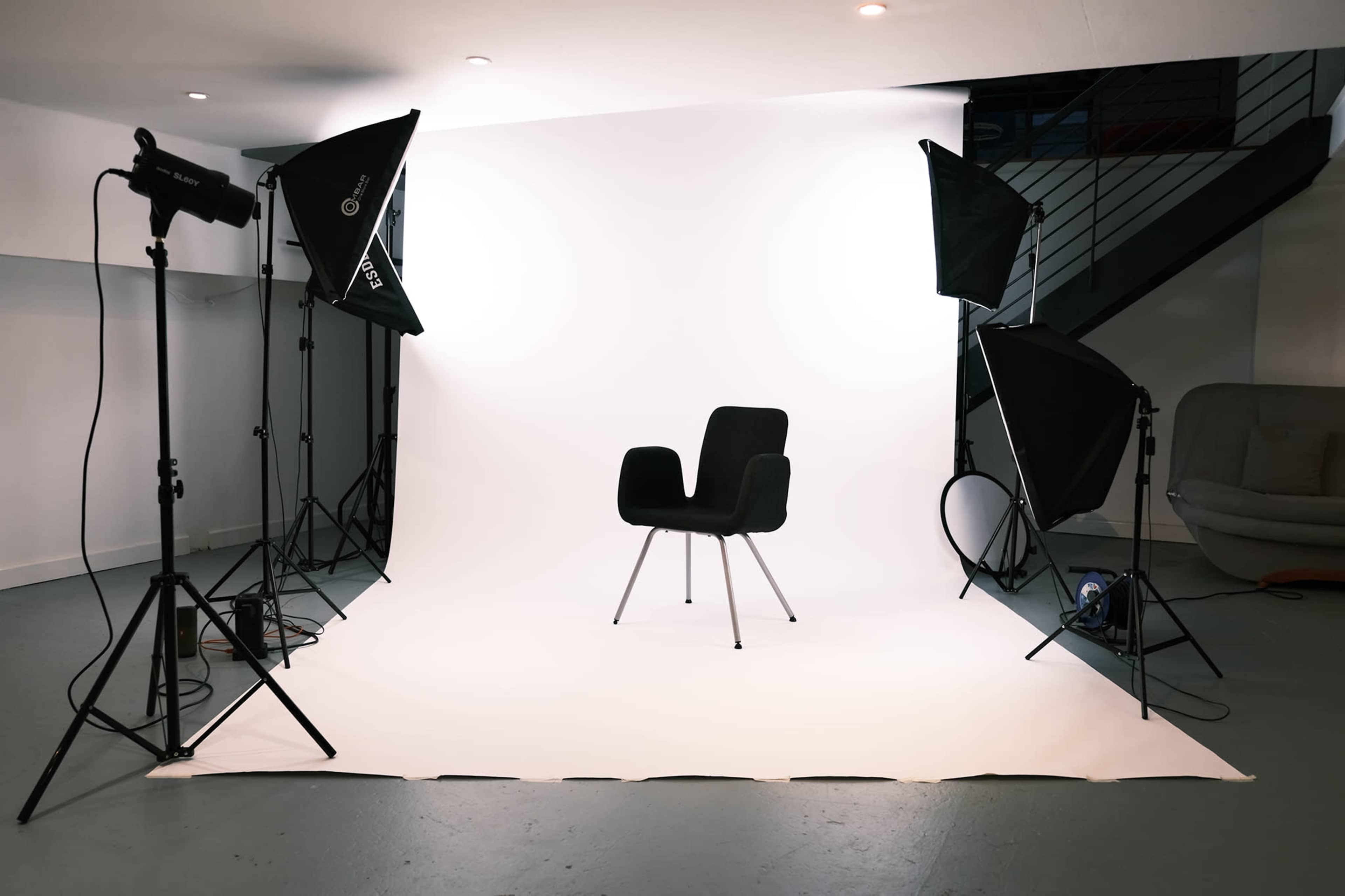 A photography studio is set up with a black chair in the center, surrounded by softbox lights and a white backdrop.