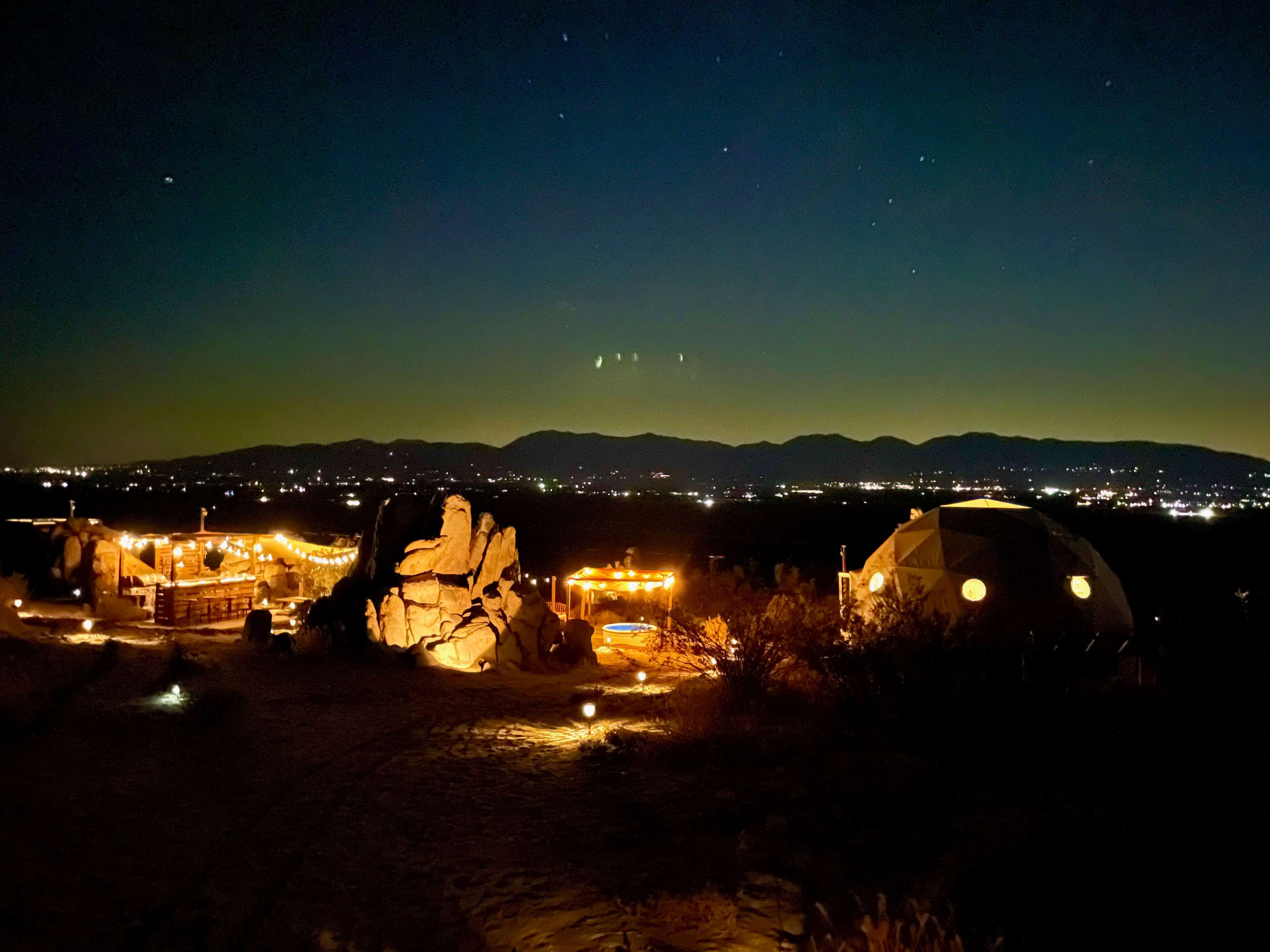 A desert landscape at night, featuring a geodesic dome and illuminated pathways among large rocks and distant mountains.