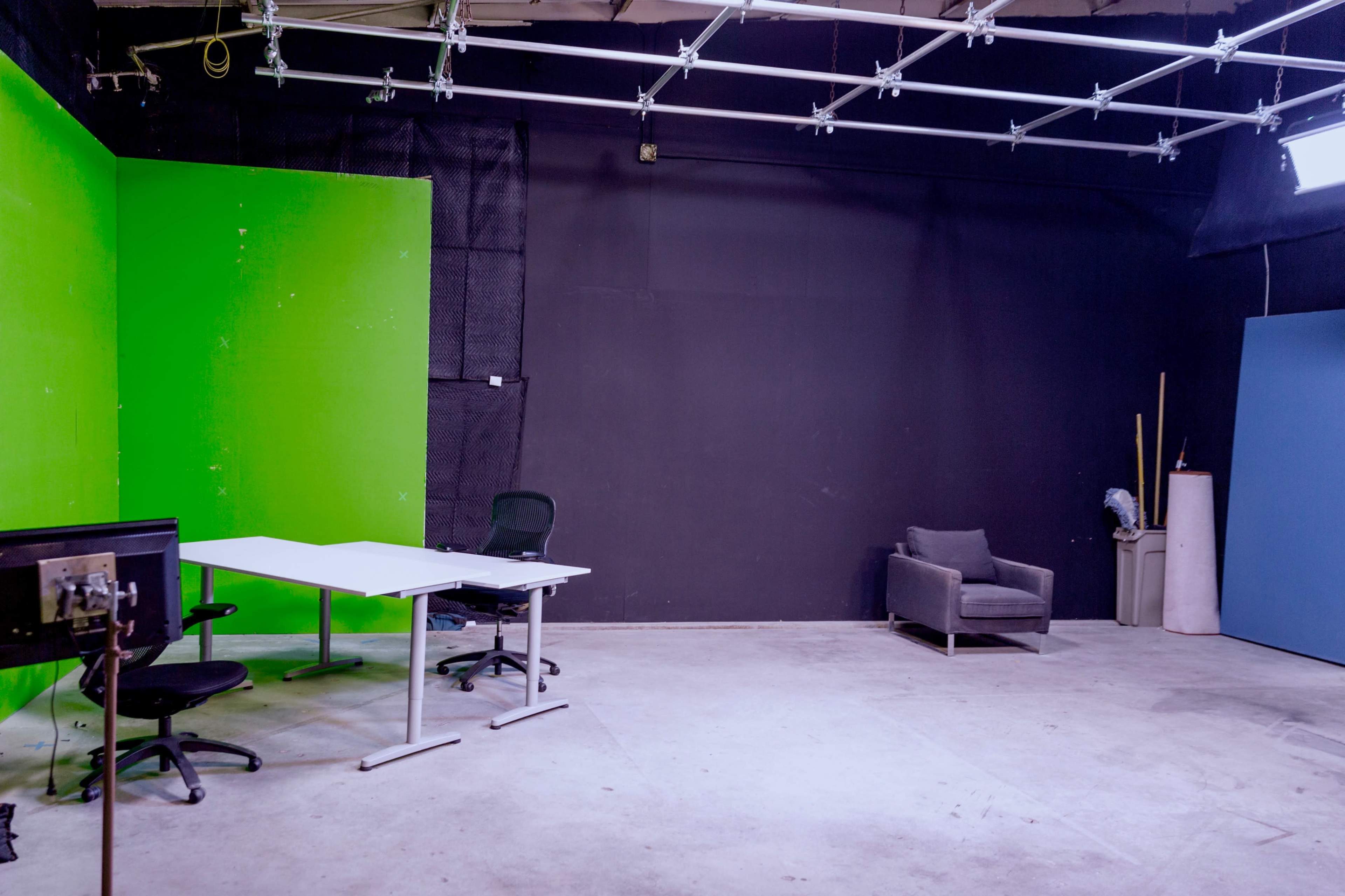 An empty studio space featuring a green screen, a black wall, a white table with chairs, and a gray couch.
