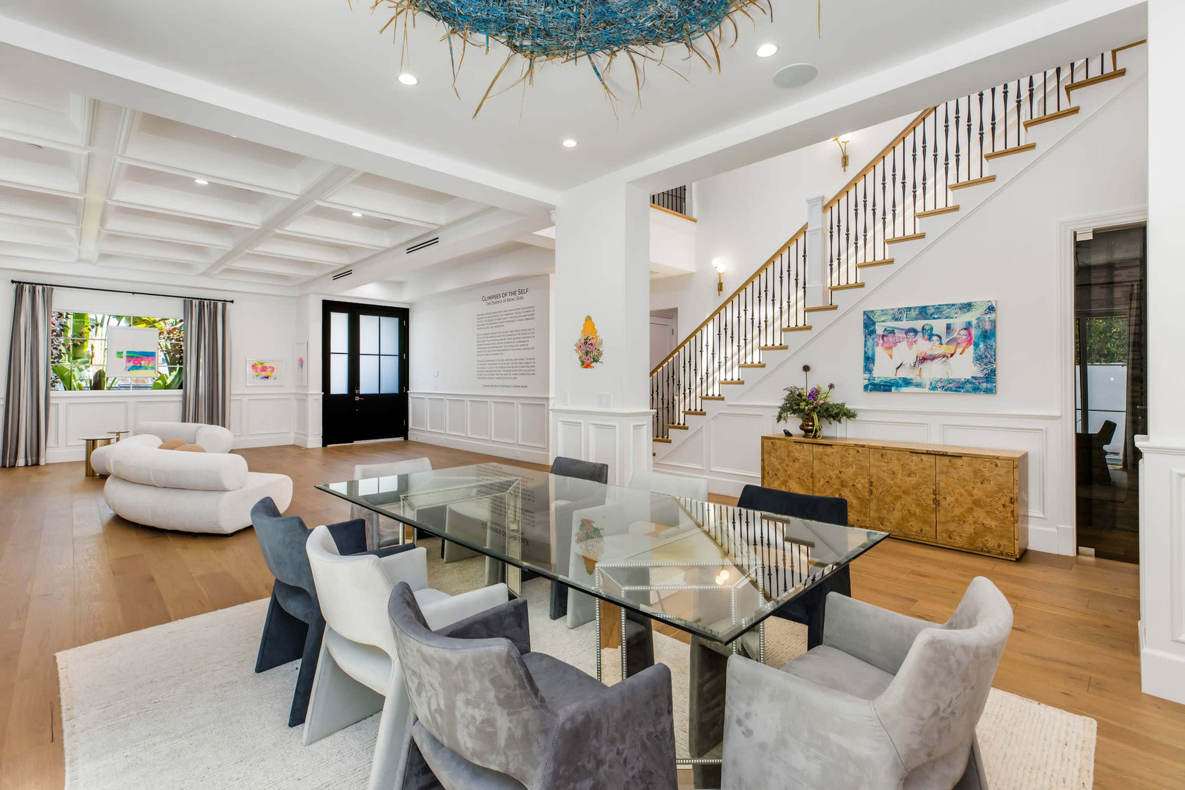The image shows a modern, open-concept dining area featuring a glass table surrounded by upholstered chairs, with a staircase in the background and large windows letting in natural light.