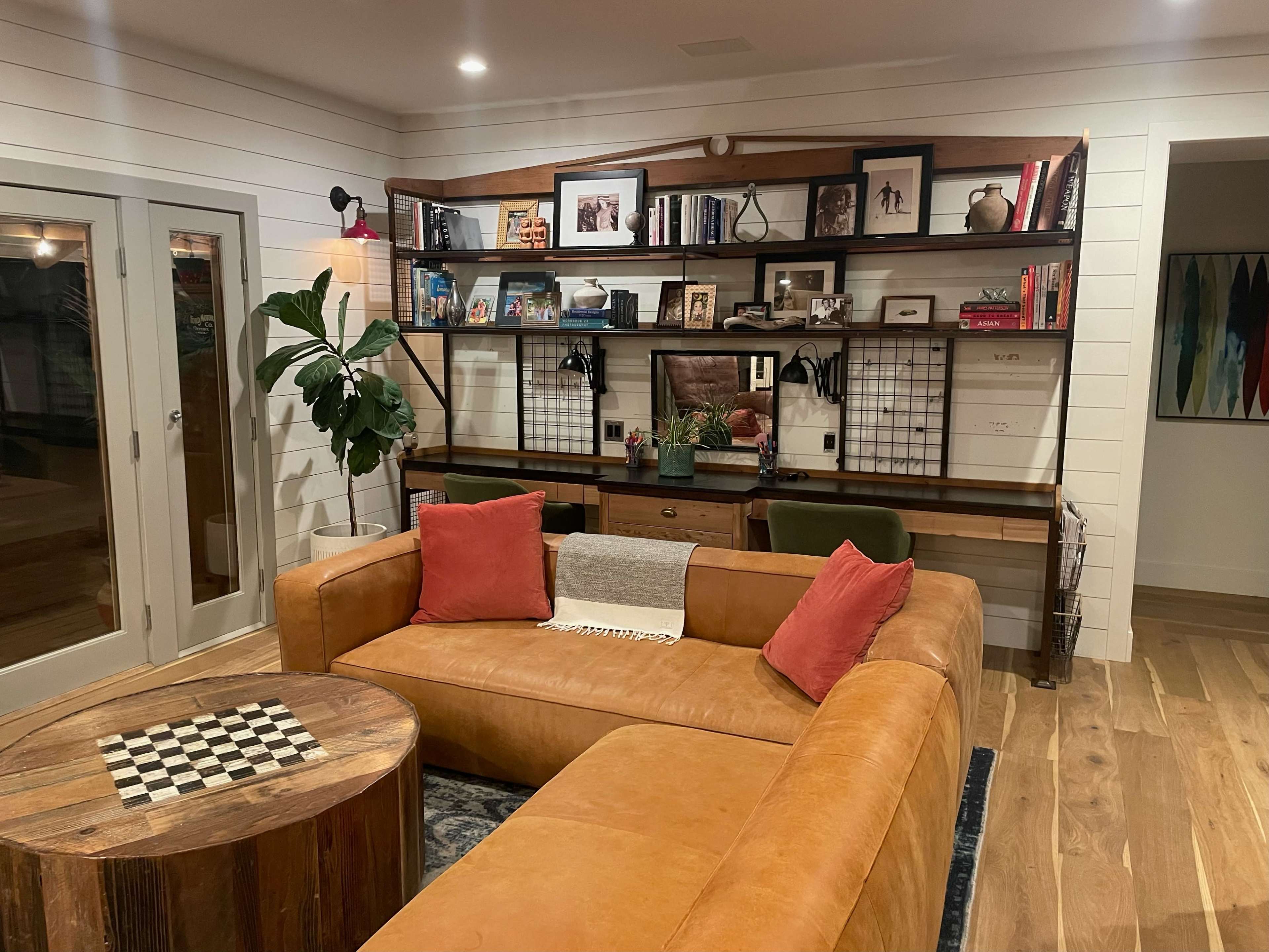 The image shows a modern living room featuring a large leather sectional couch, a wooden coffee table with a checkered top, and a wall shelf filled with books and decorative items.