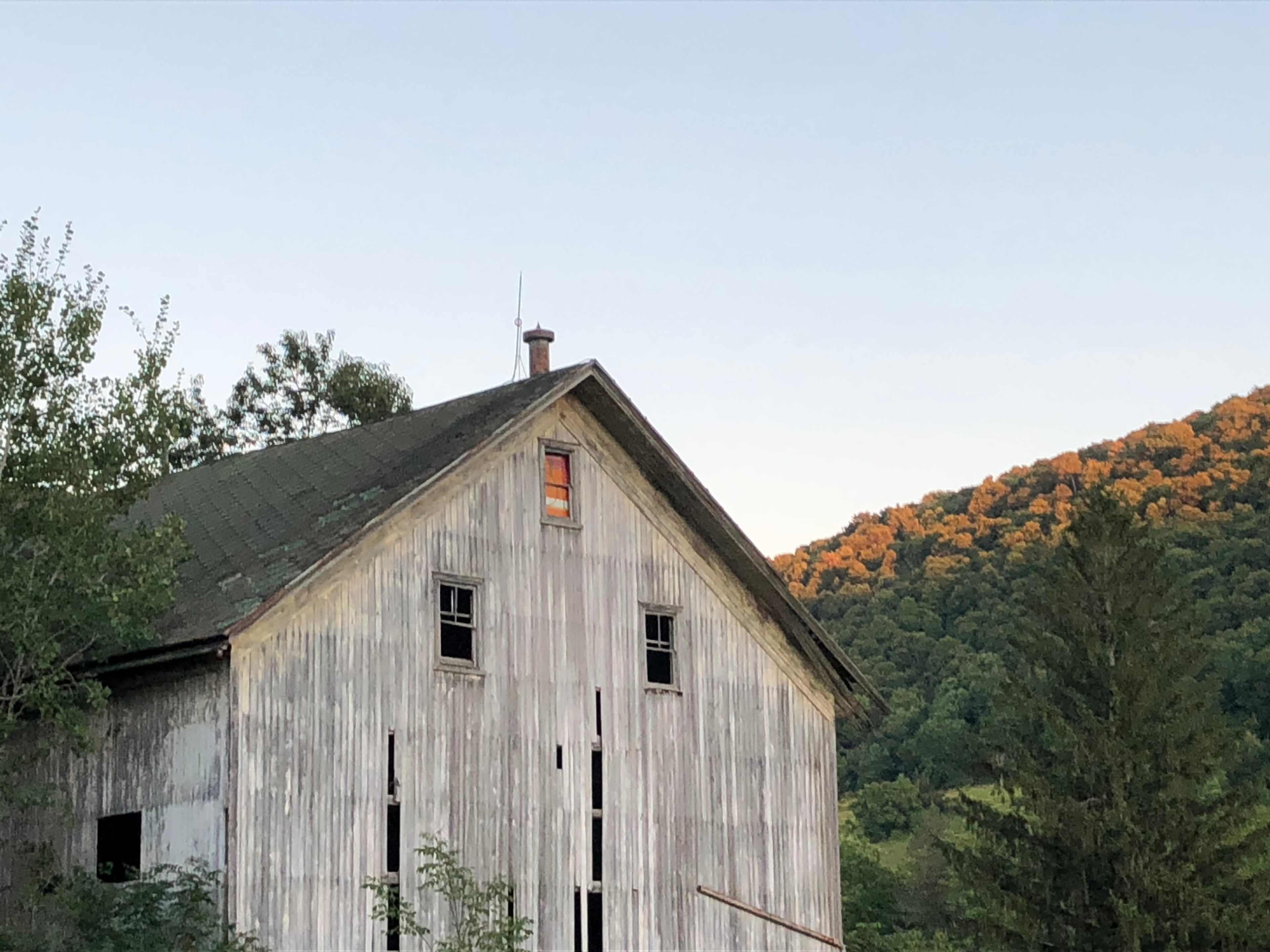 A weathered barn stands against a backdrop of wooded hills with autumn-colored foliage.