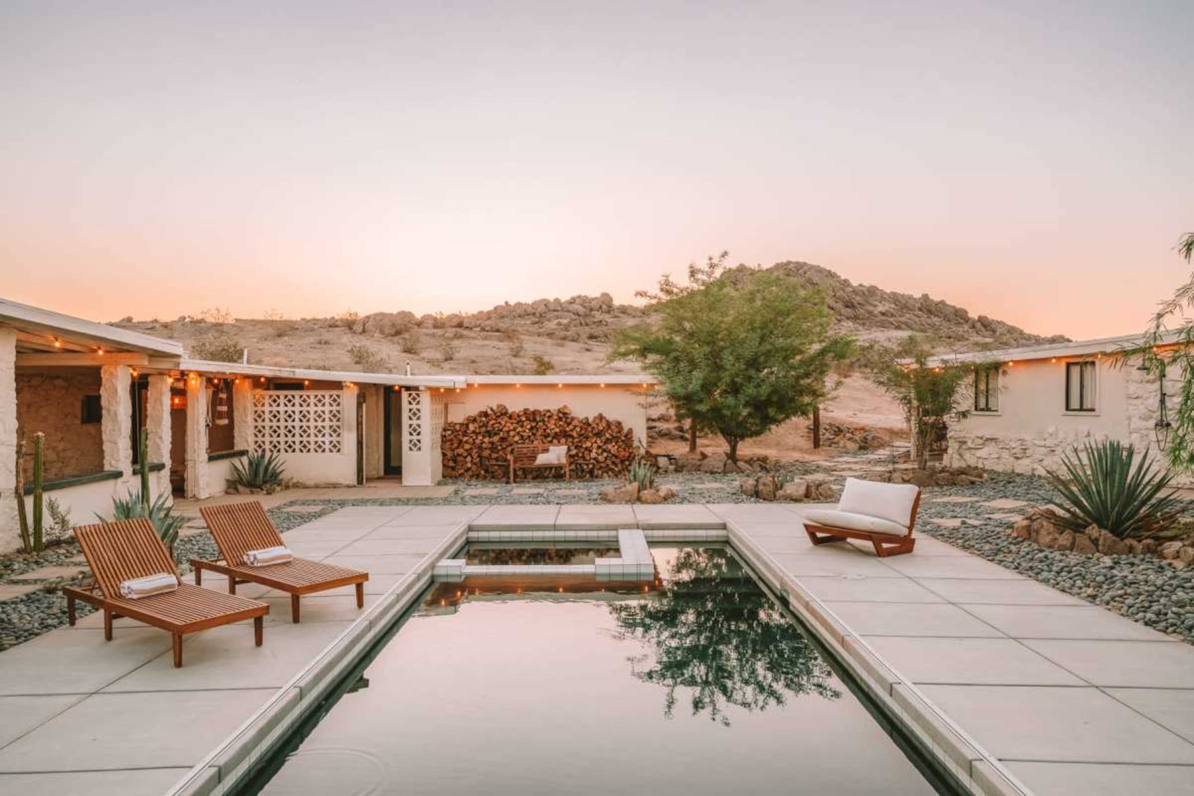 The image shows a modern desert retreat featuring a swimming pool, wooden lounge chairs, and landscaping with rocks and plants, set against a mountainous backdrop at sunset.