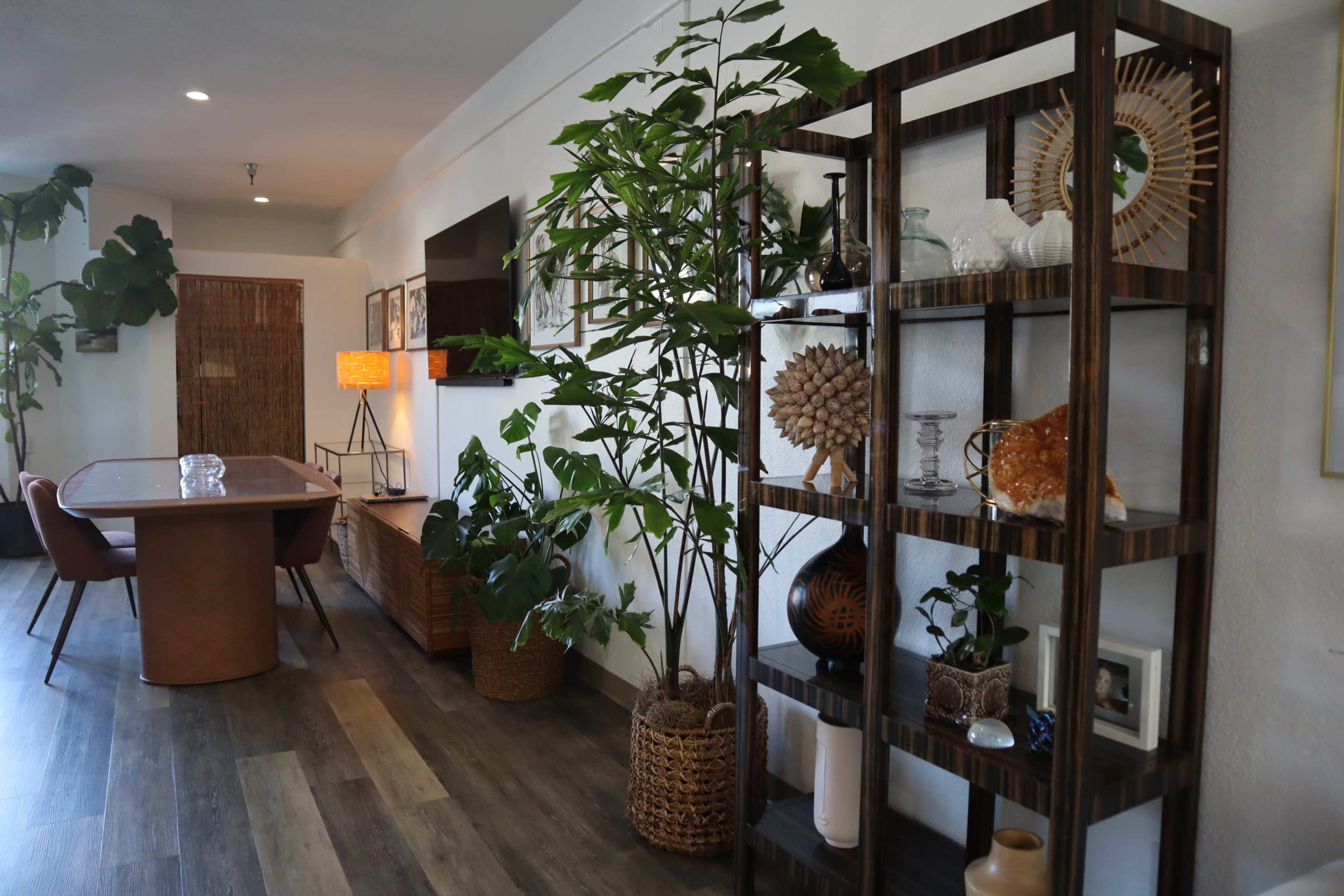 A modern interior space featuring a dining table, a decorative shelving unit with plants and ornaments, and a warm-toned lamp.