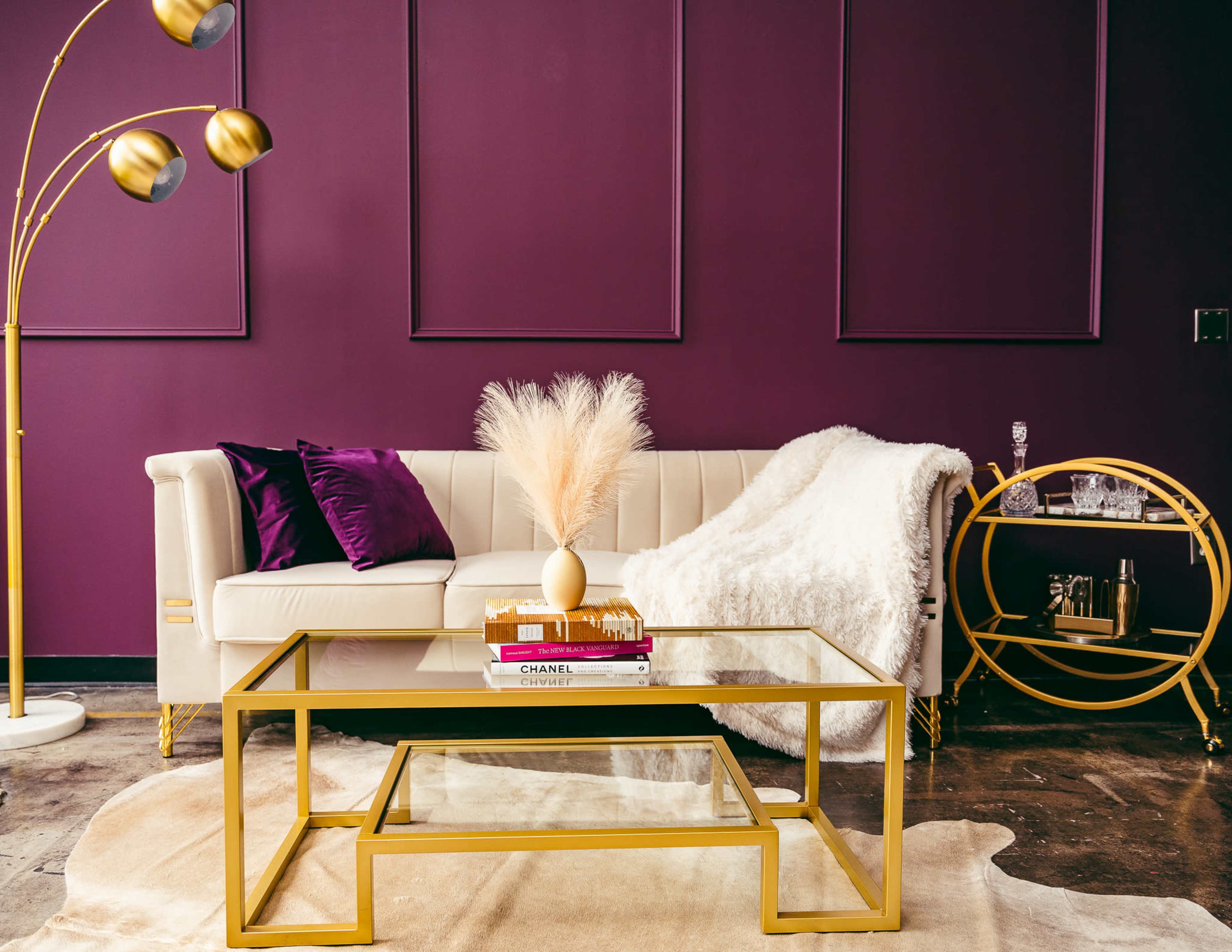 A white sectional sofa with purple cushions is arranged in front of a gold coffee table, accompanied by a floor lamp, decorative vase, and a side table, set against a deep purple wall.