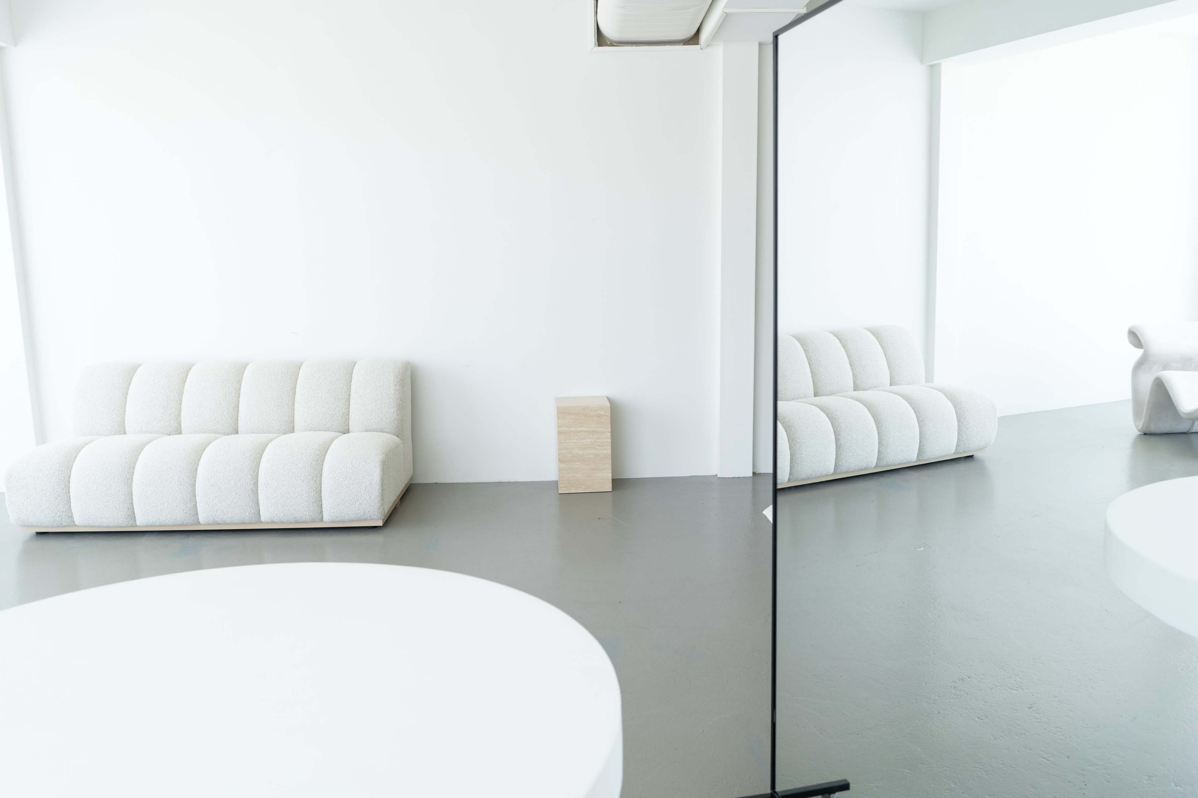 The image shows a minimalistic interior with two light-colored sofas and a wooden side table, reflected in a large mirror on the left.