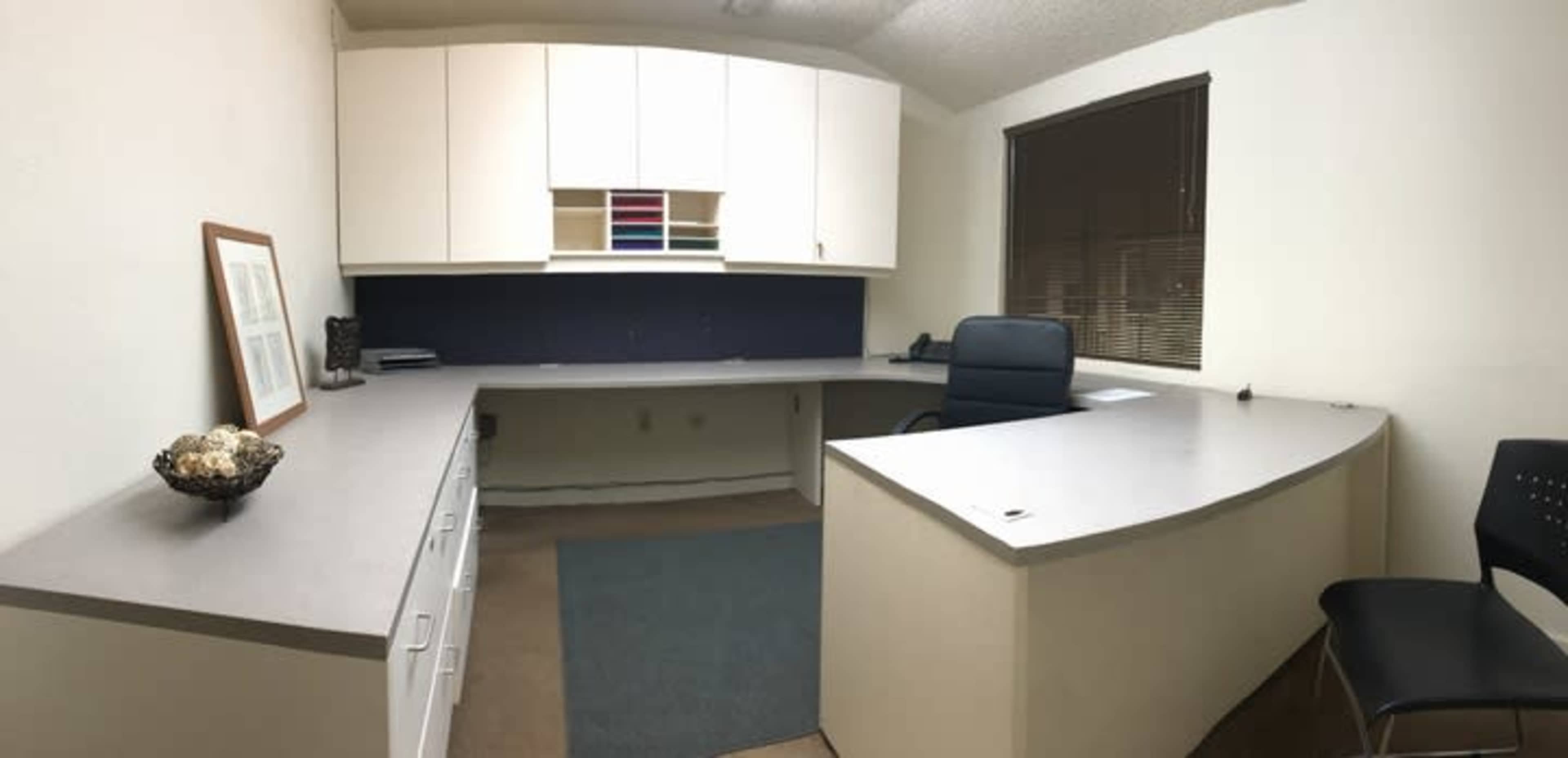 An office space featuring a large L-shaped desk, a black office chair, and a decorative bowl on a table, with shelves above the desk and window blinds.