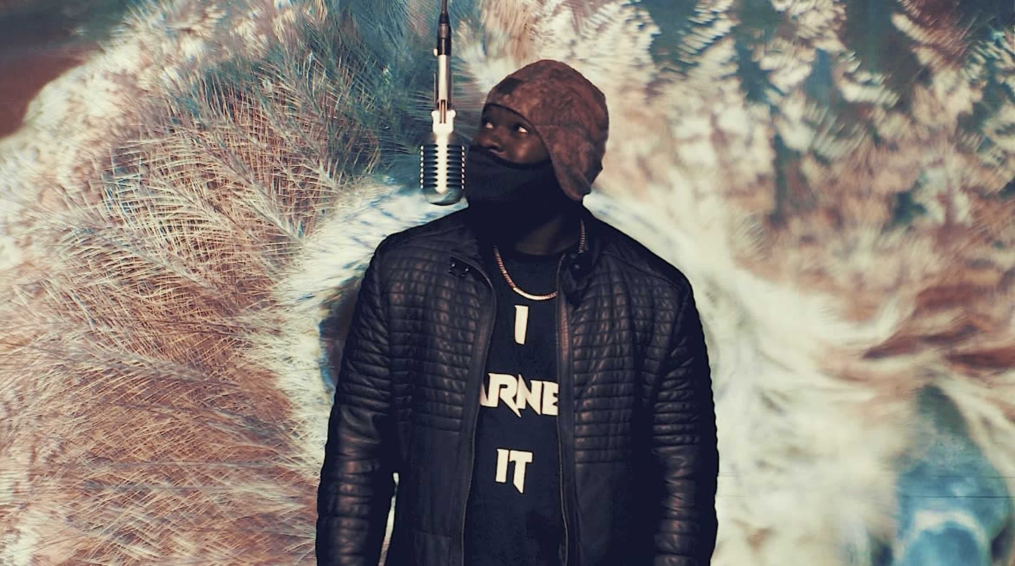 A person in a hat and mask stands in front of a colorful, patterned backdrop with a microphone above.