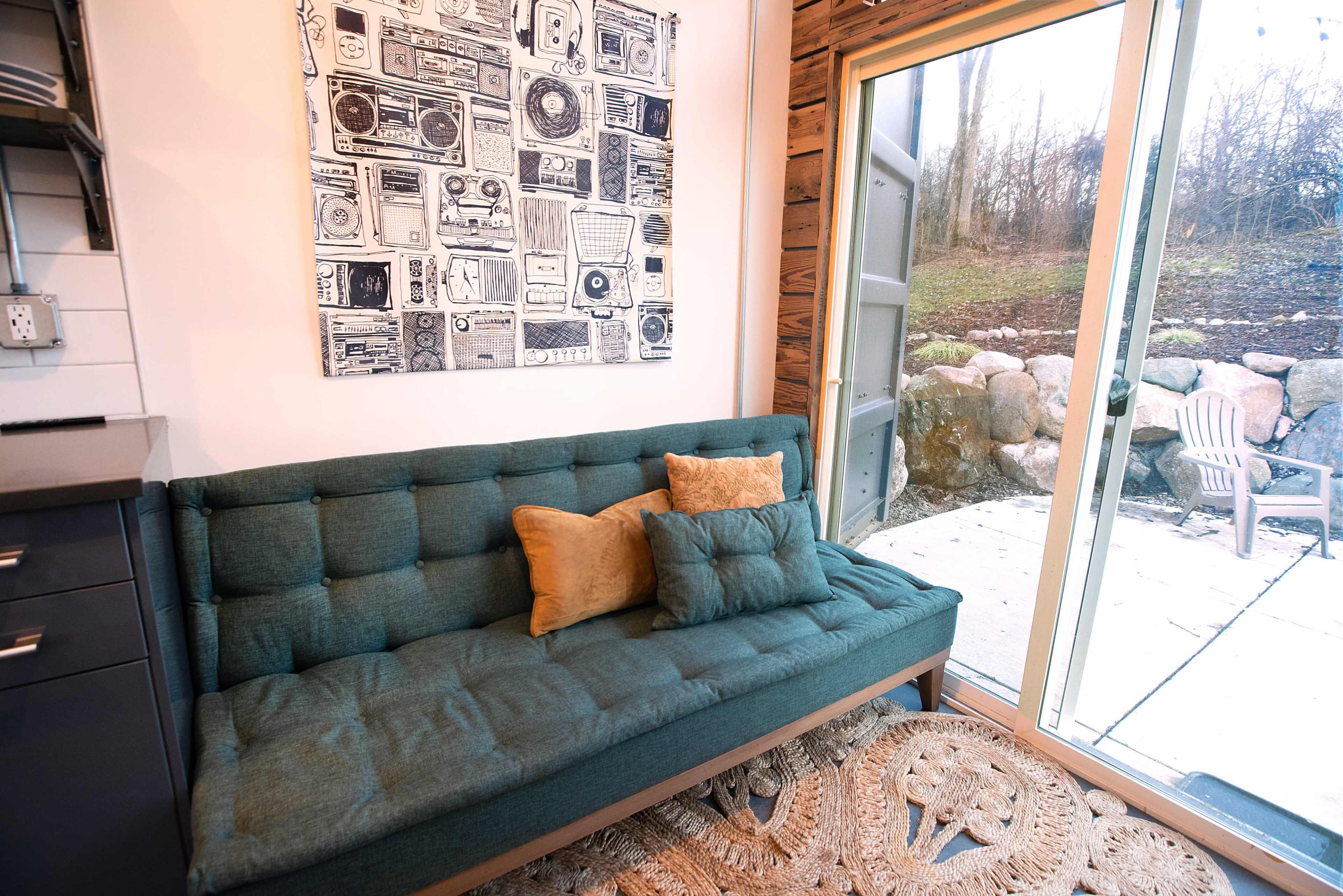 A teal sofa with decorative pillows is positioned near a large glass door that opens to a stone patio and a wooded area.