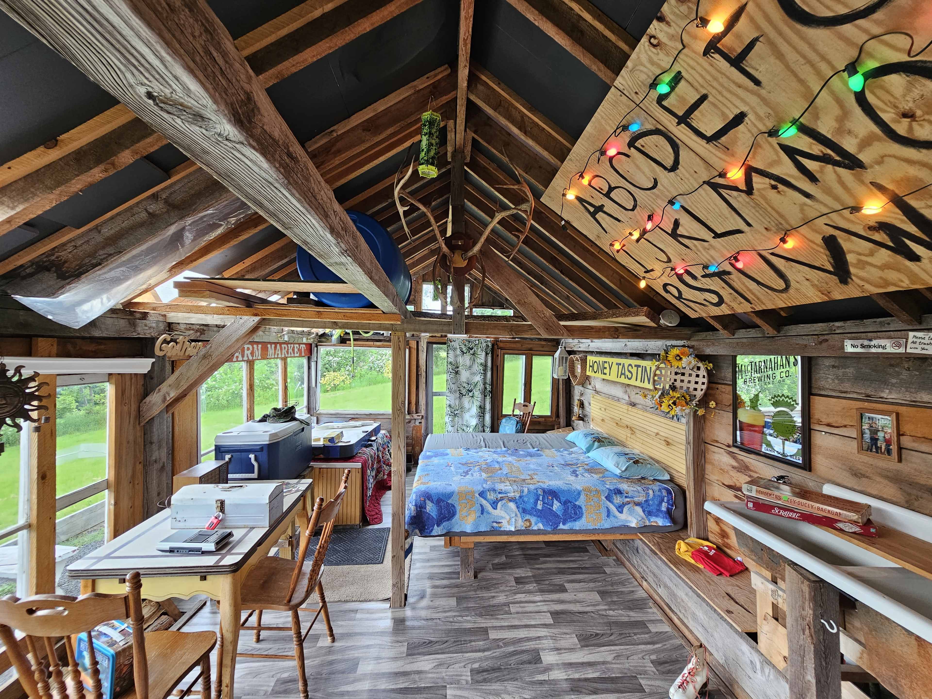 The image shows a rustic interior of a wooden cabin with a bed, a dining area, and colorful string lights adorning the walls.