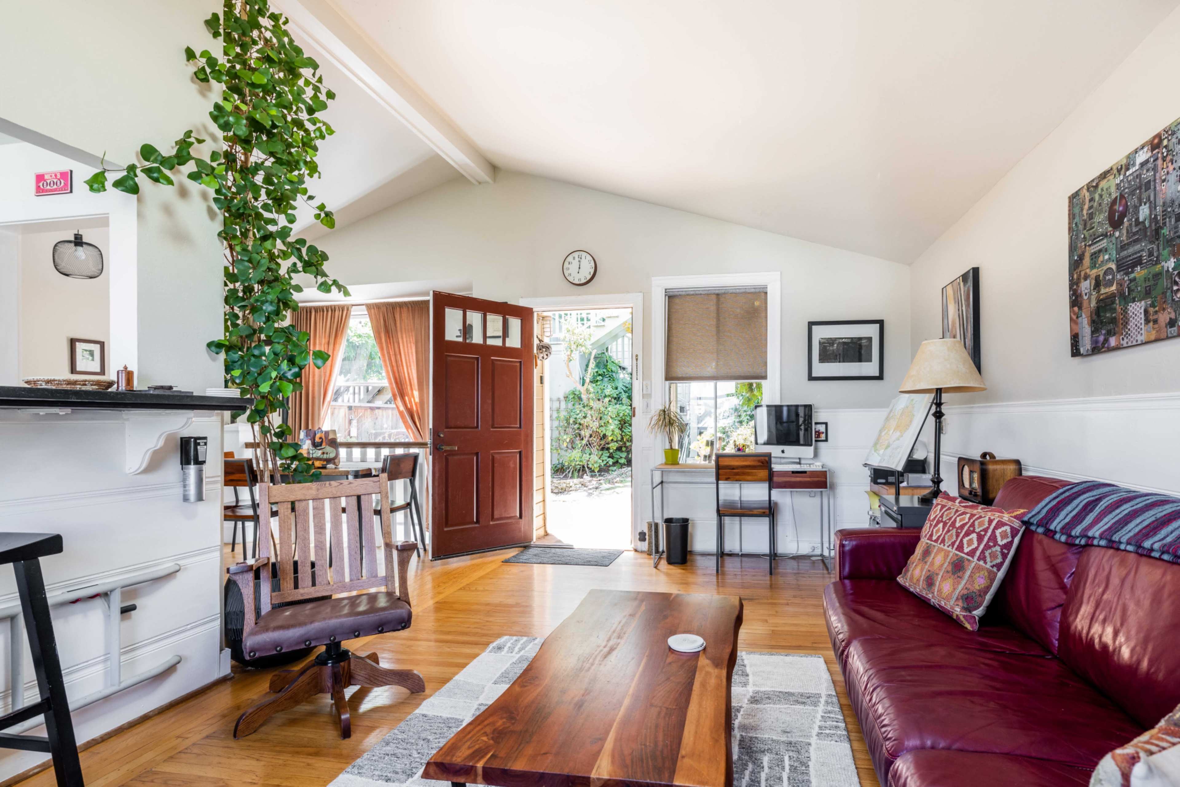 The image shows a cozy living room with a red couch, a wooden coffee table, and an open front door leading to a bright outdoor space.