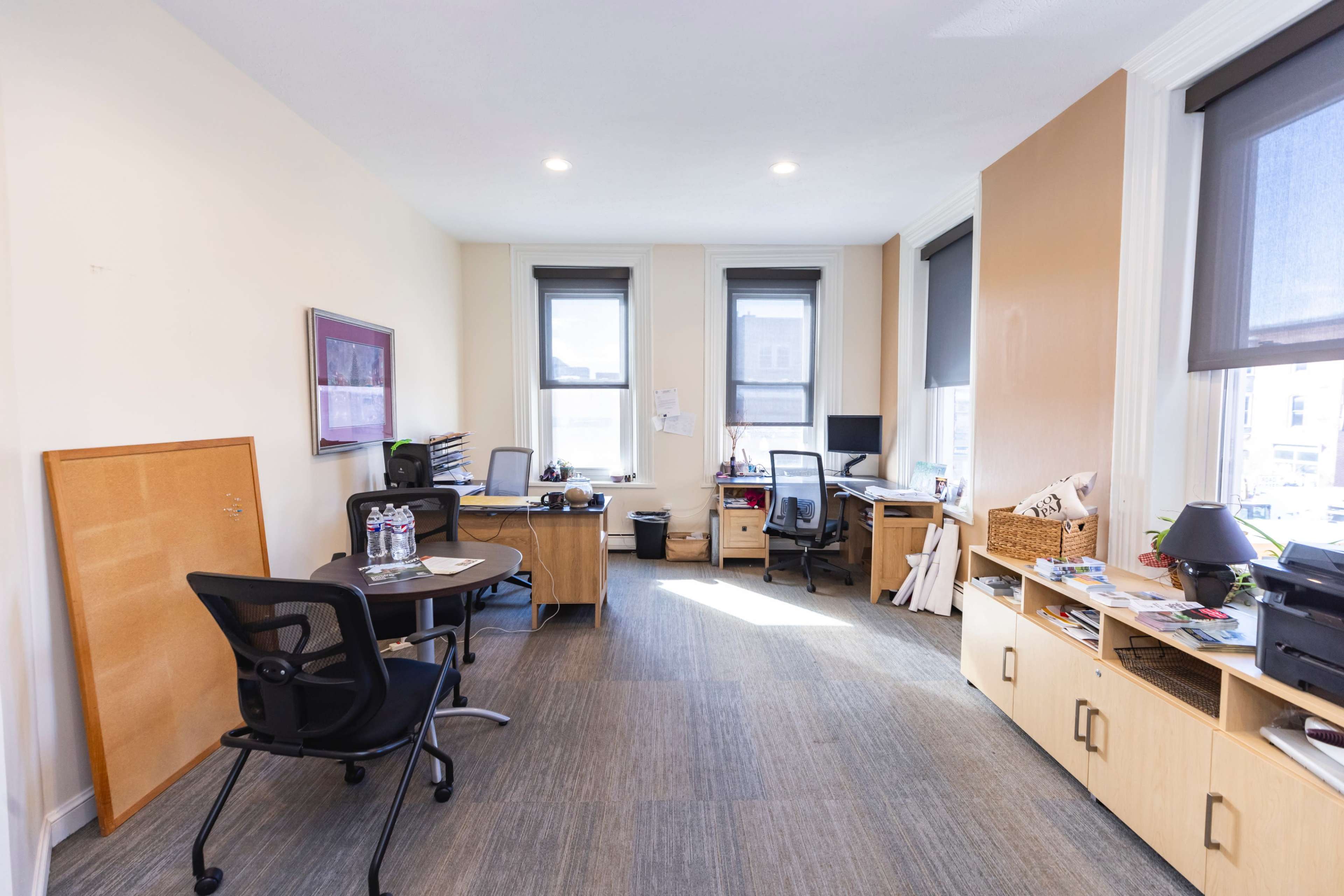 An office space with two desks, chairs, and various office supplies arranged in a well-lit room with large windows.