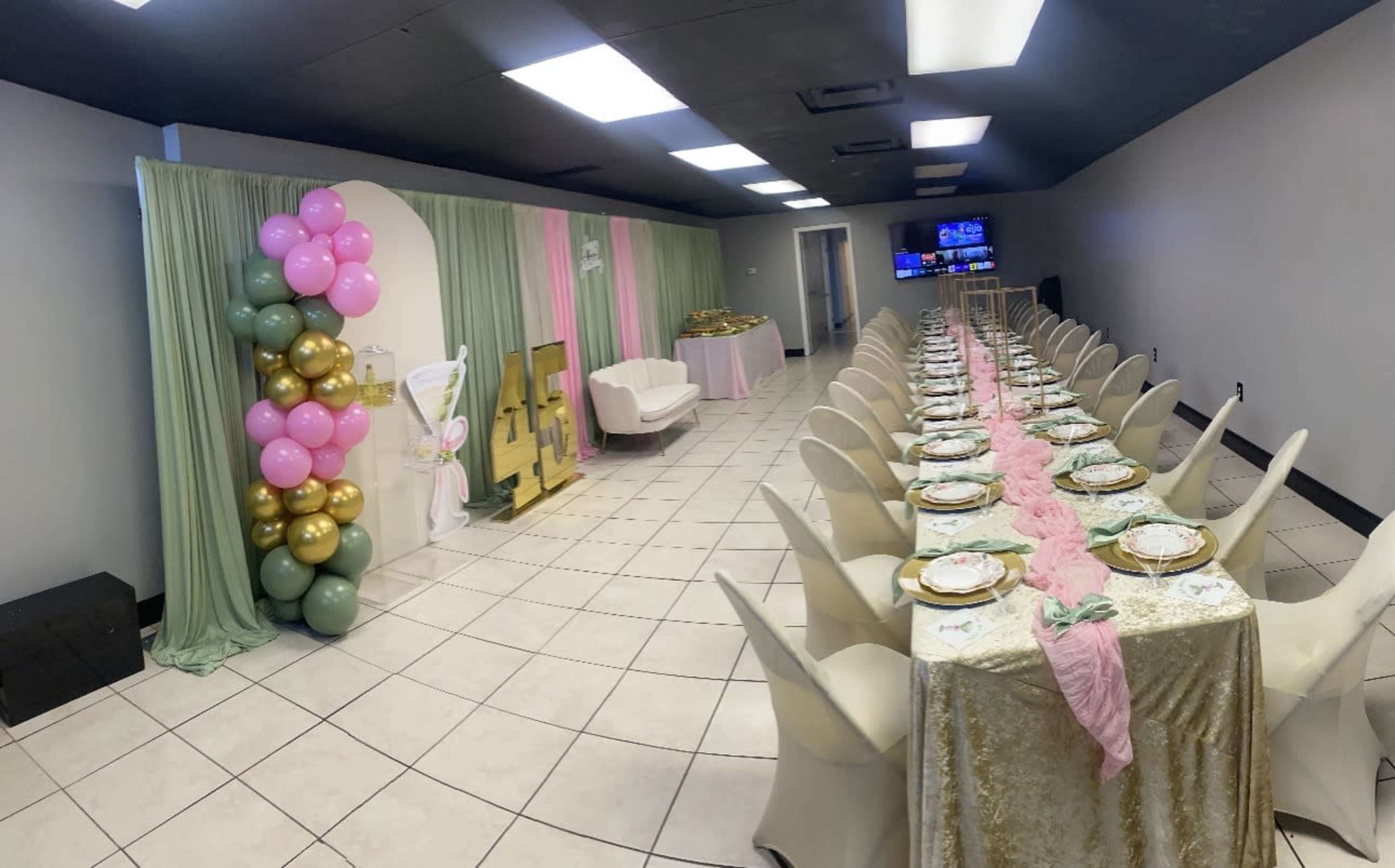 The image shows a decorated event space with a long dining table set with plates and pink runner, surrounded by pastel balloons and a backdrop featuring a "Happy Birthday" sign.