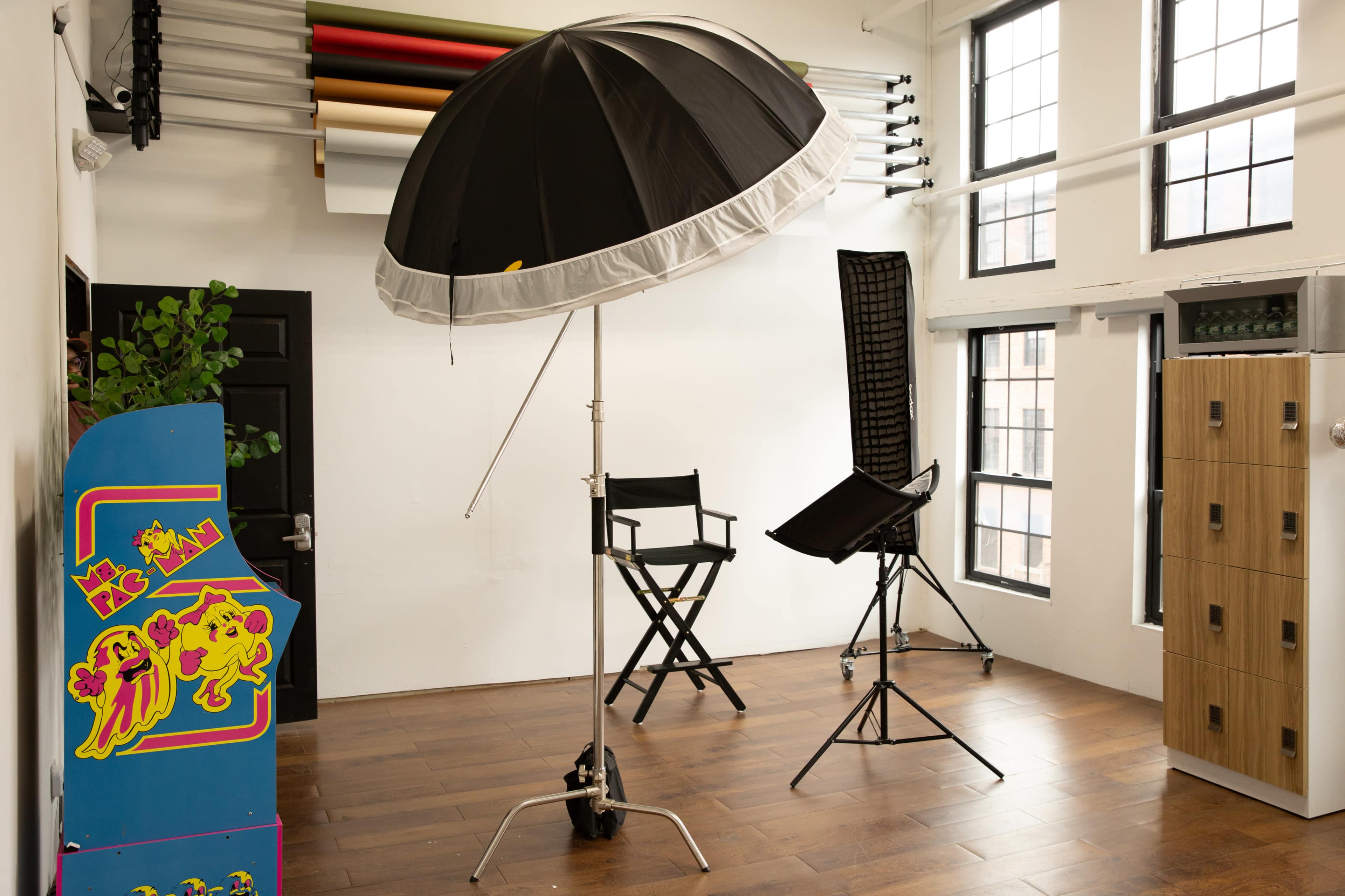 The image features a photography studio with a large black and white umbrella light, a director's chair, a tall black seat, and wooden lockers on the side, illuminated by natural light from large windows.