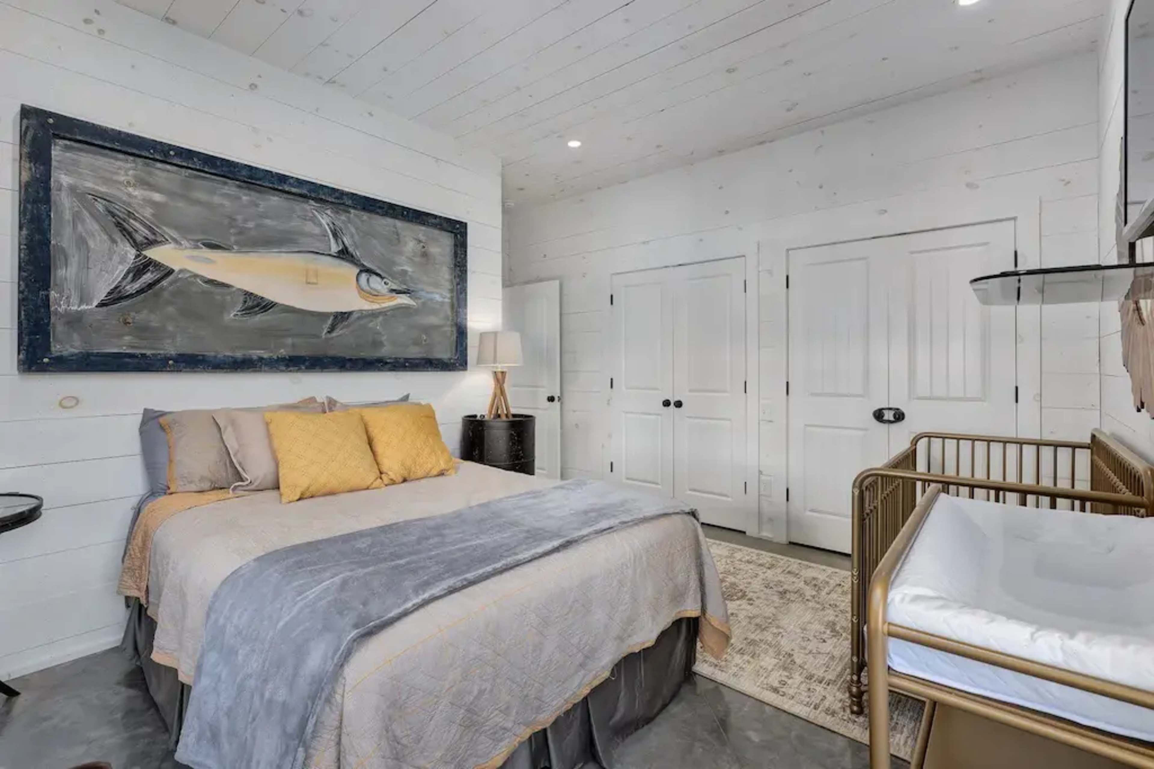 A bedroom featuring a bed with gray and yellow bedding, a large fish painting on the wall, and a nearby baby crib.