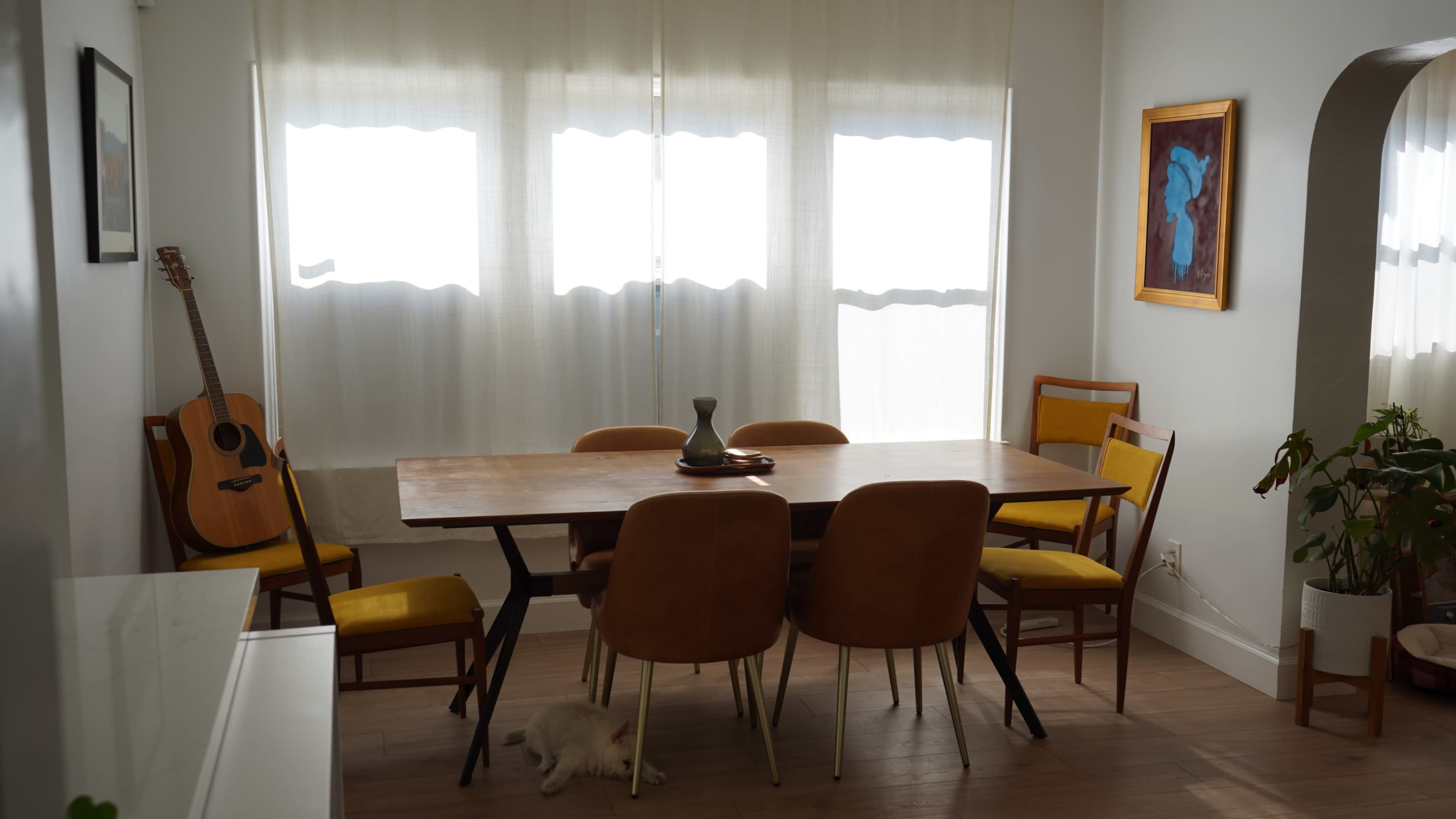 A dining room features a large wooden table surrounded by six chairs, with a guitar leaning against the wall and a potted plant nearby.