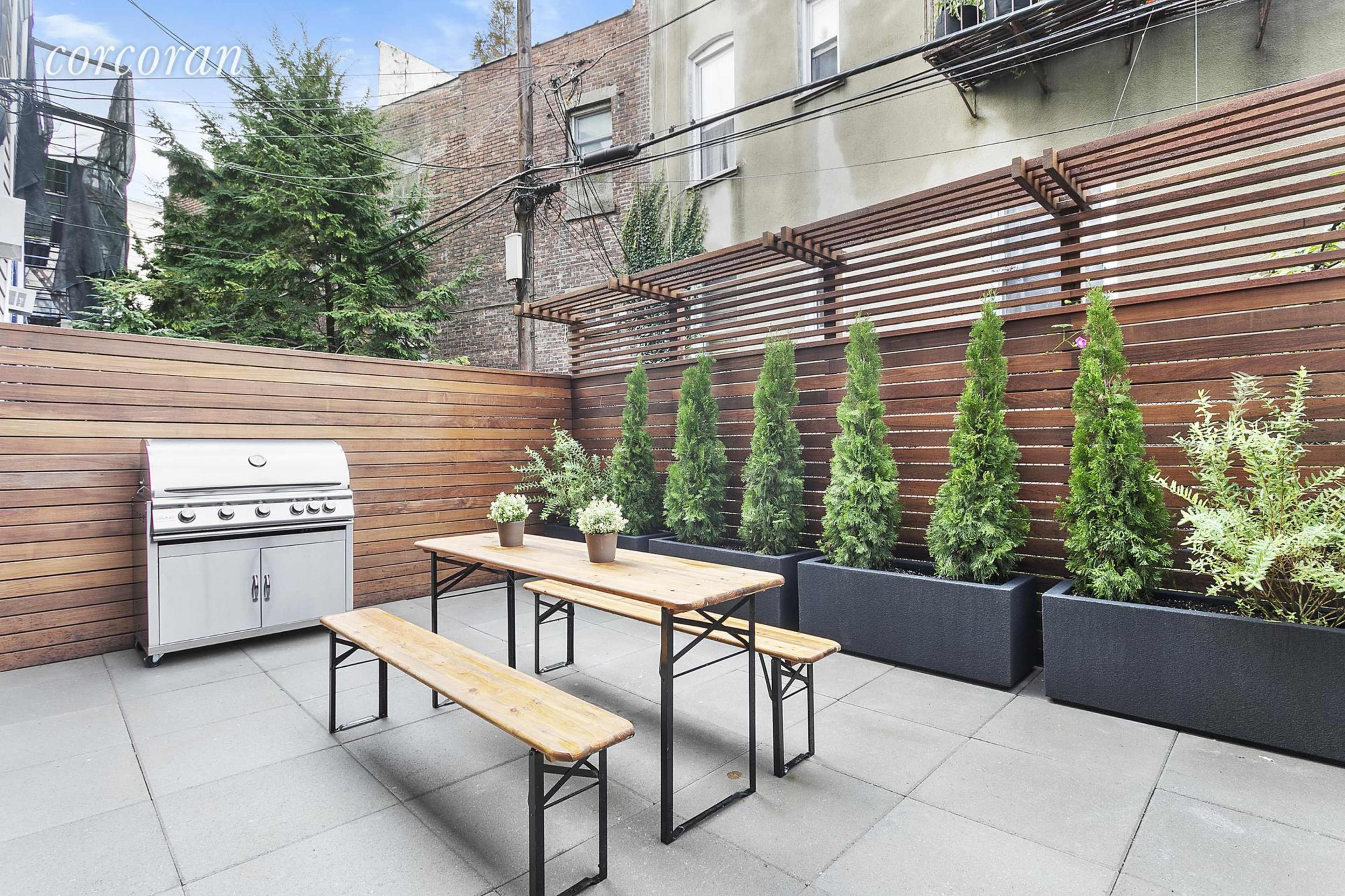 A backyard patio featuring a stainless steel grill, a wooden table with benches, and neatly arranged planters with tall green shrubs.
