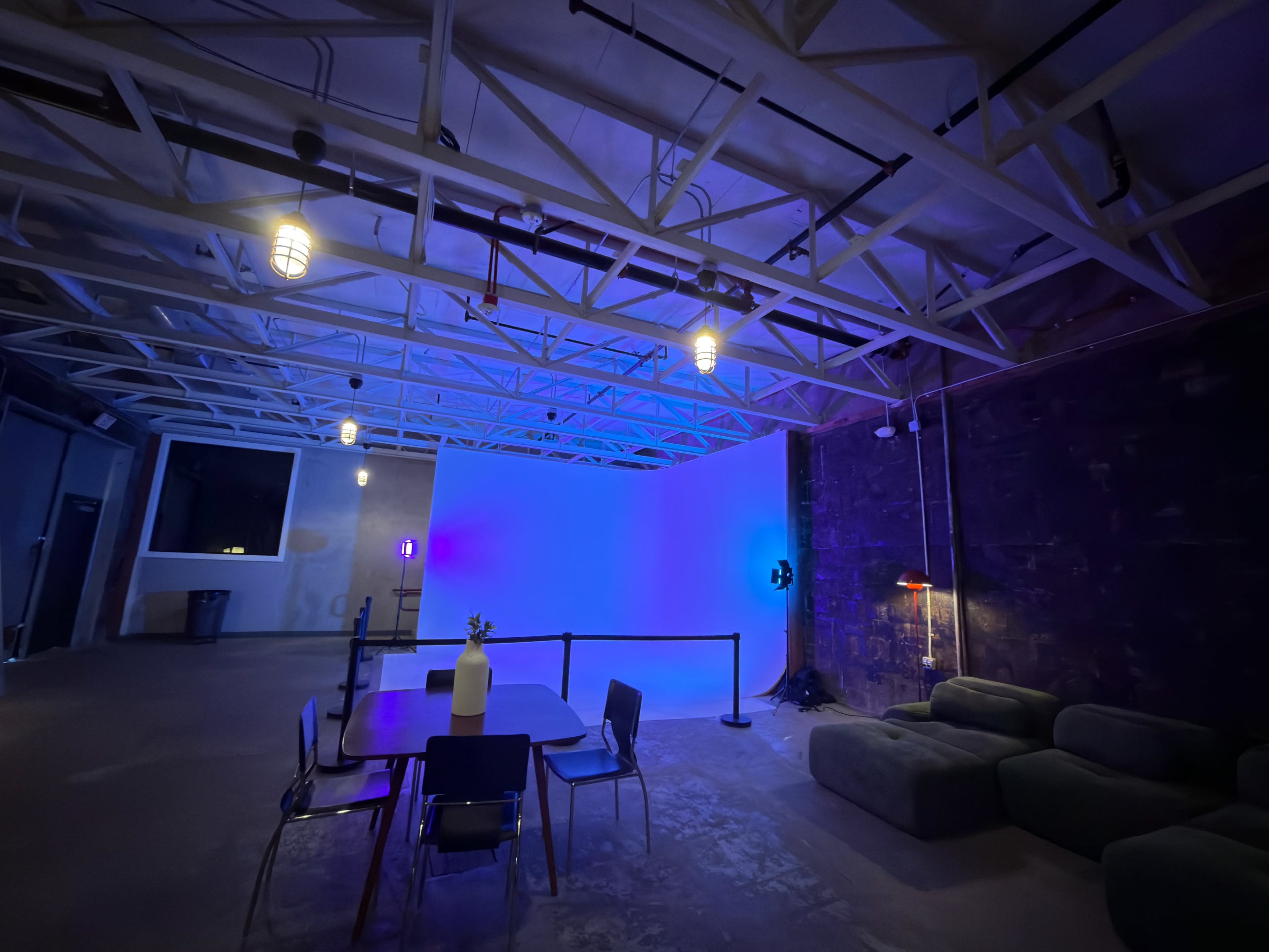 The image shows a spacious, modern room with a large table and chairs, a couch against the wall, and various colorful lights illuminating the area.