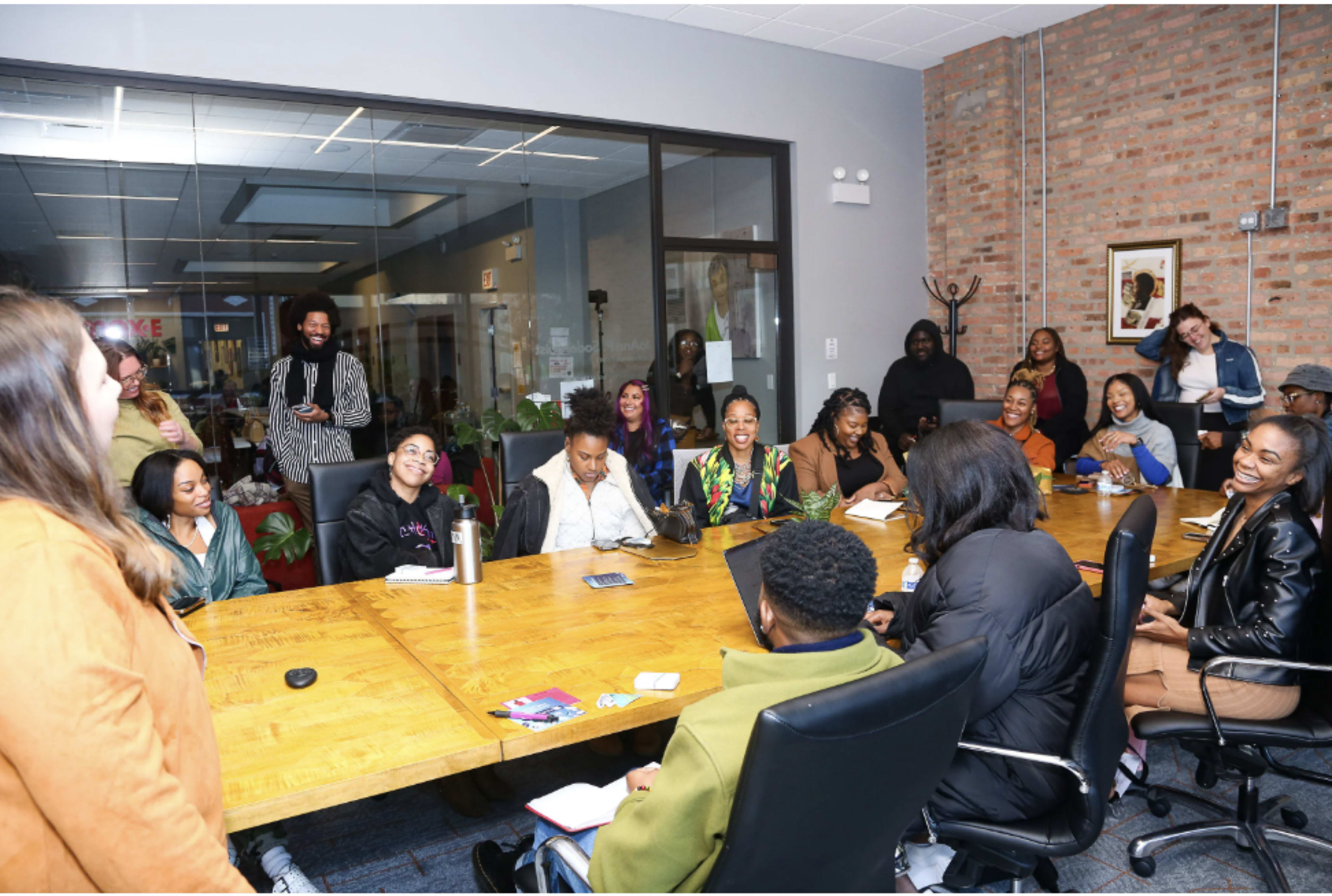 A diverse group of people are seated around a large wooden table in a brightly lit meeting room, engaging in conversation and interaction.