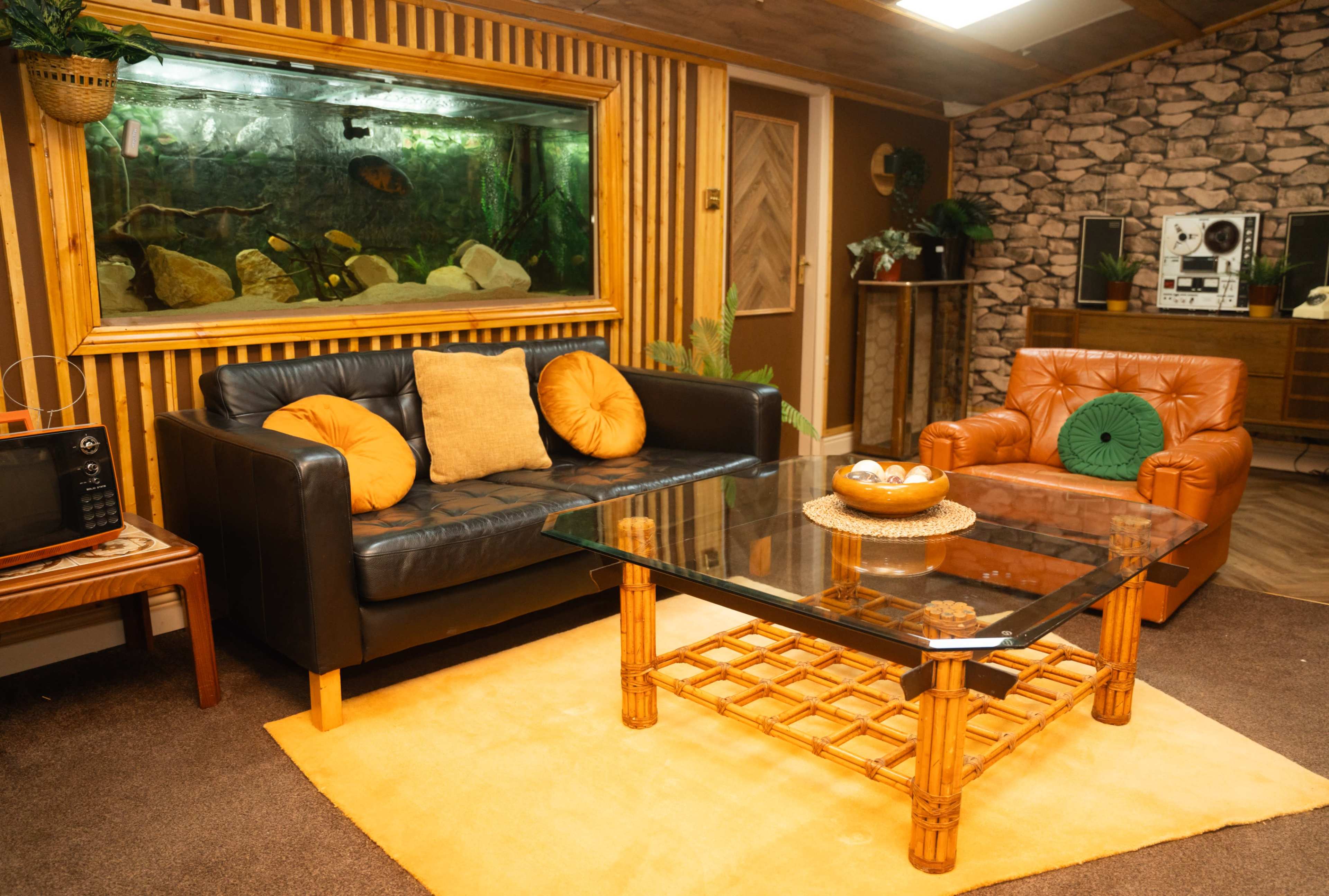 The living room features a black leather sofa, a brown armchair, a glass coffee table with a rattan base, and a large aquarium set against a wooden wall.