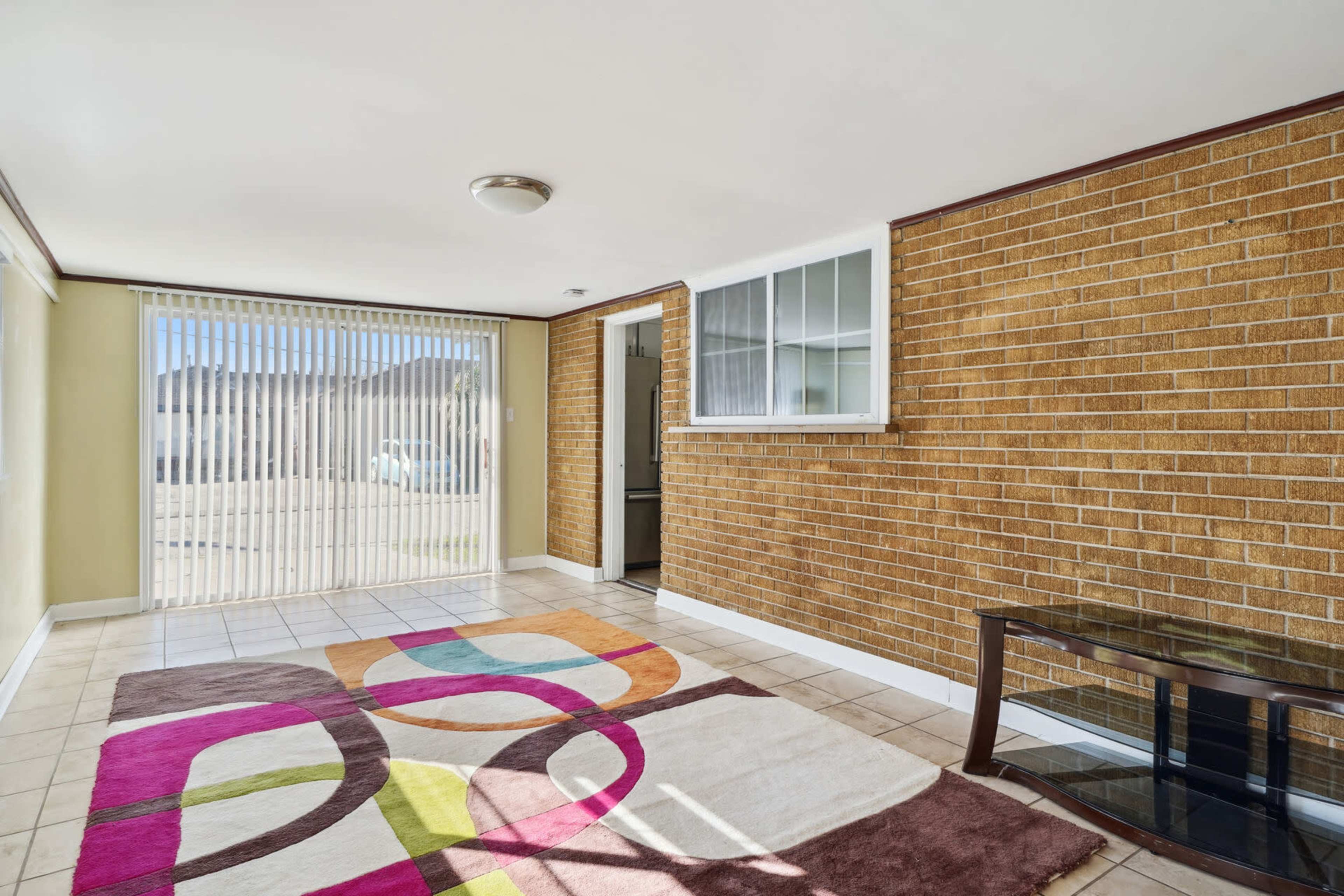 The image shows a bright, spacious room with a tiled floor, a colorful area rug, and a large window covered by vertical blinds.