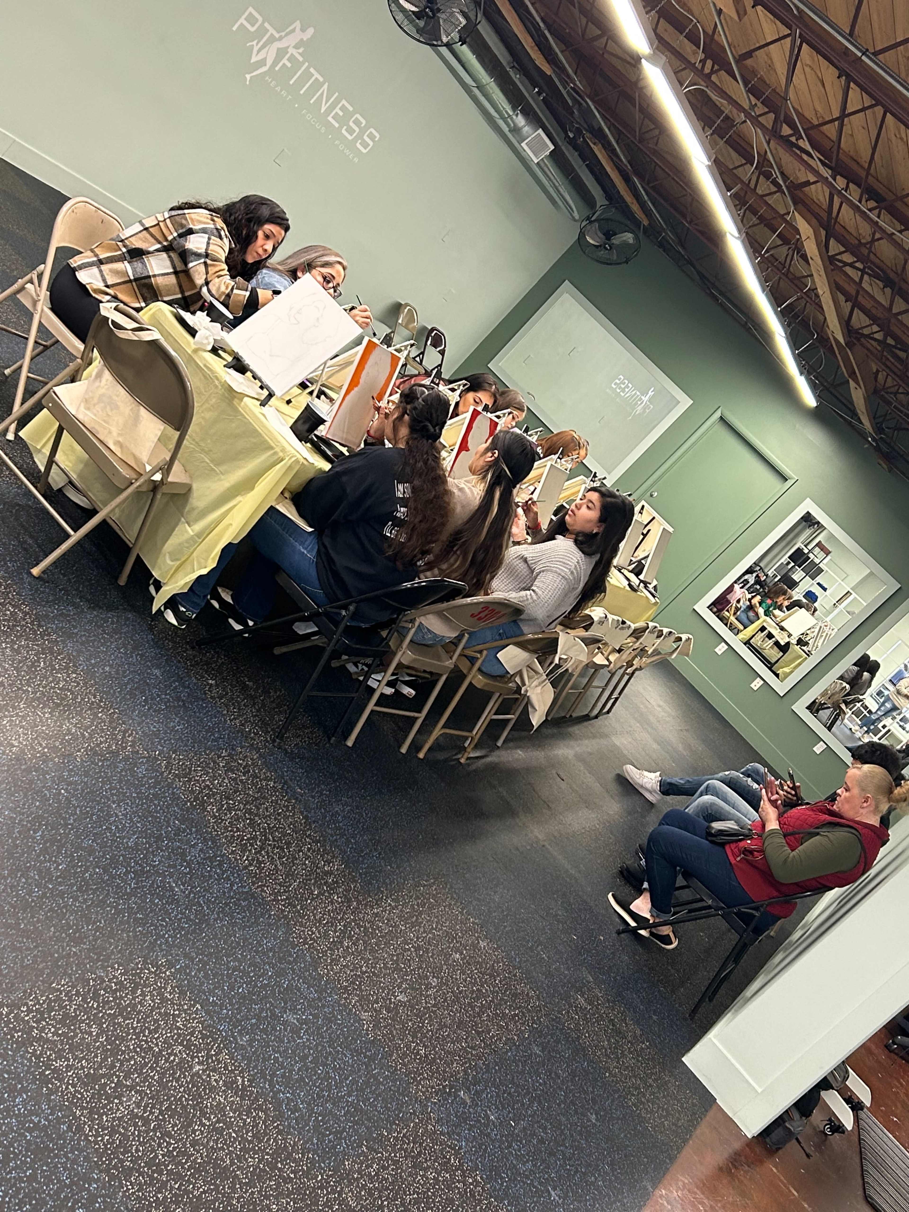 A group of people is seated at tables in a fitness studio, engaged in an art or craft activity.