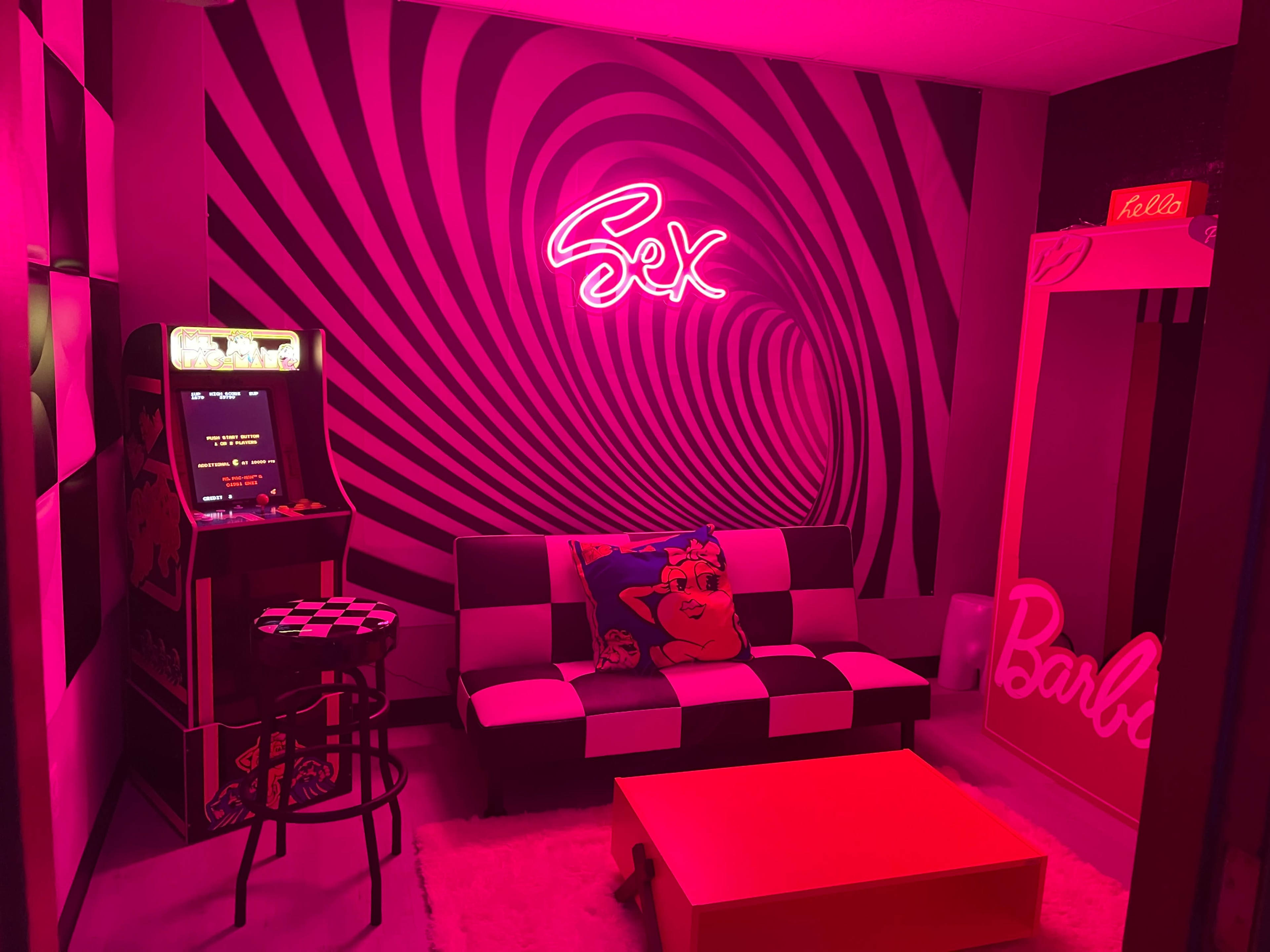 The room features a black-and-white checkered sofa, a neon sign reading "Sex," a vintage arcade game, and a mirror with the word "Barbie" on the side.