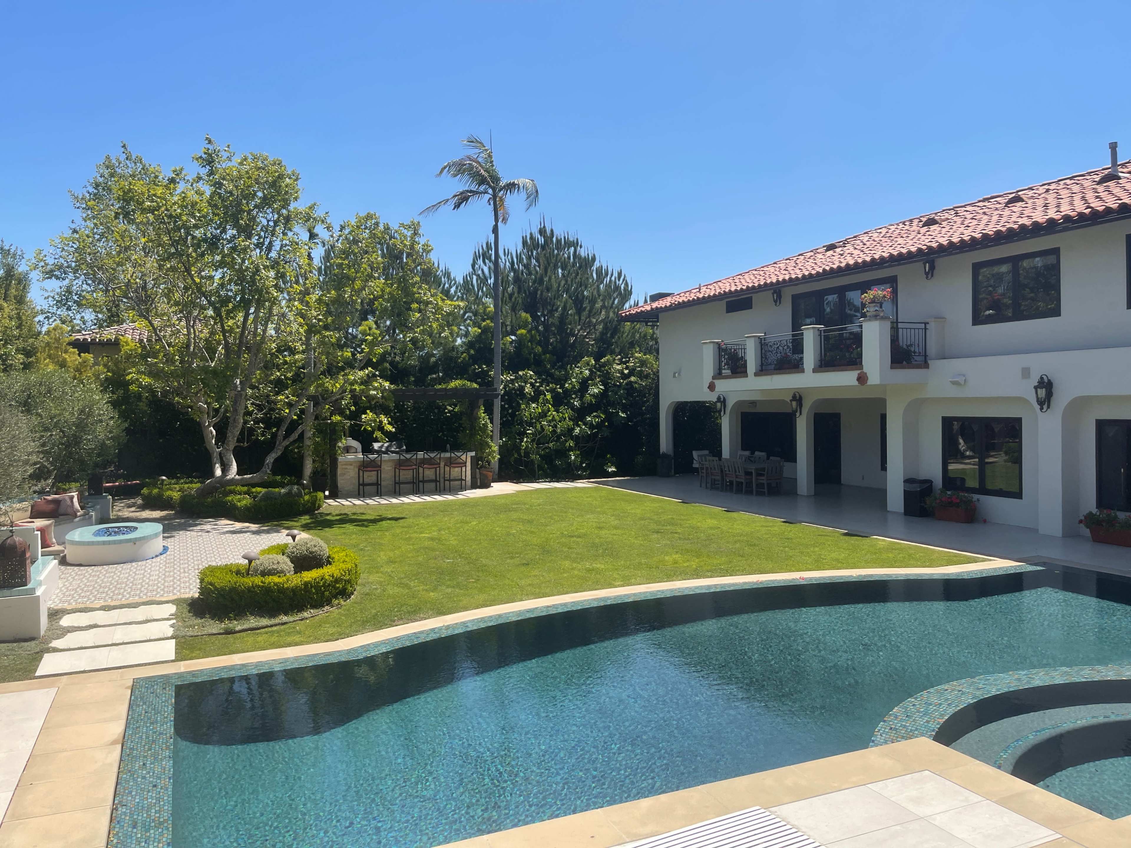 A luxurious backyard with a swimming pool, neatly manicured lawn, and a dining area surrounded by trees and greenery.