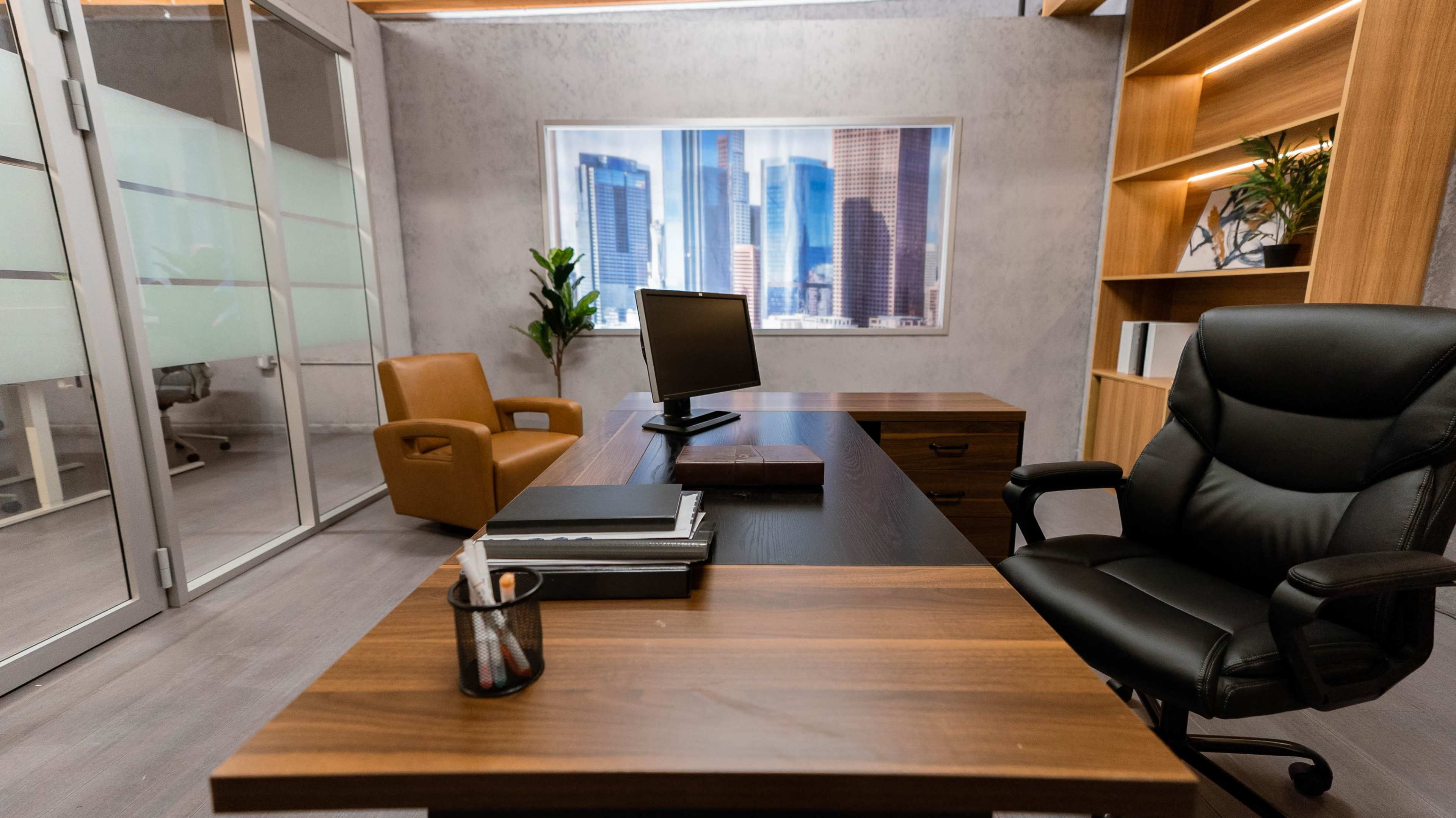 A modern office space features a wooden desk, a swivel chair, and a view of city skyscrapers through a large window.