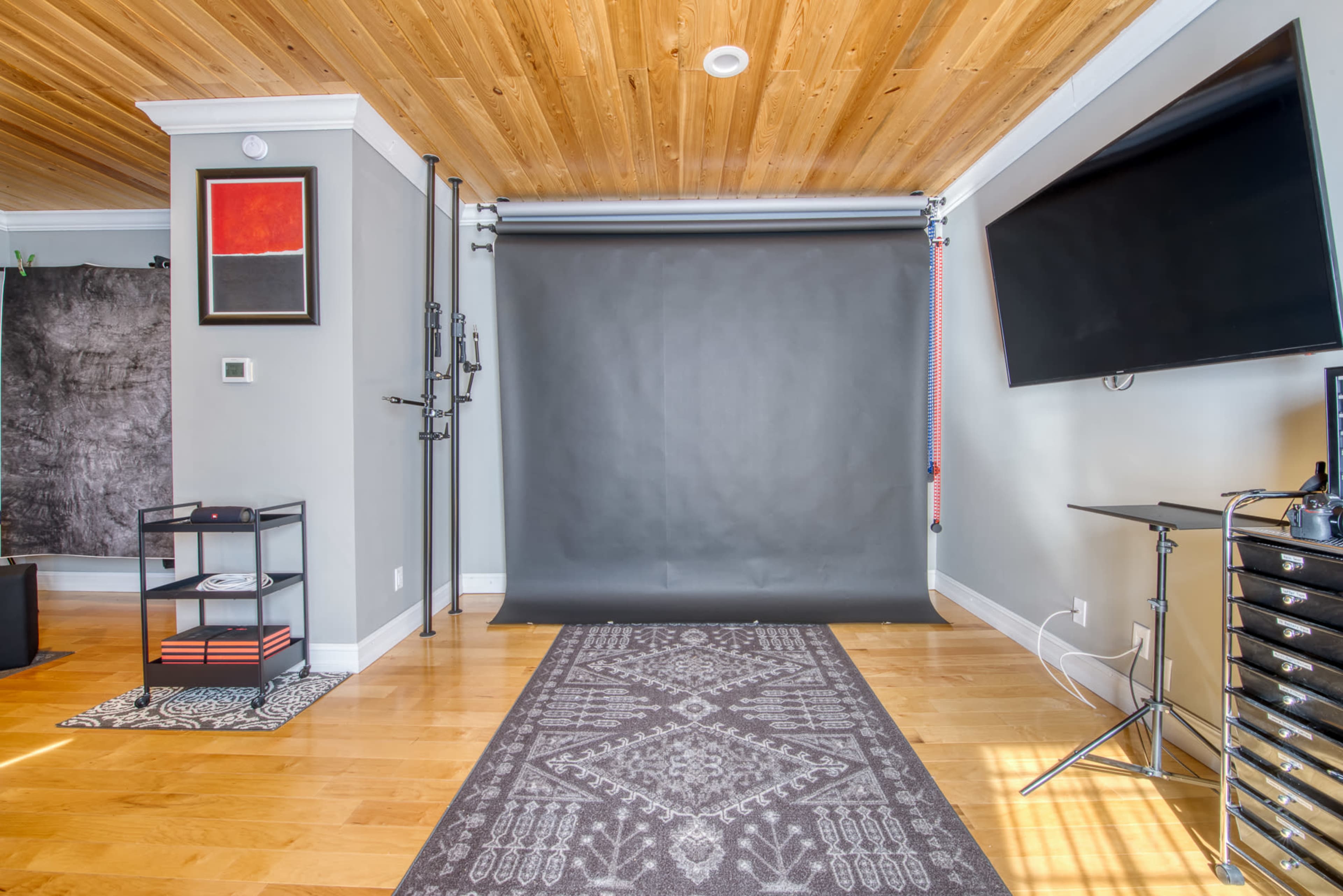 A spacious room with a wooden ceiling, featuring a dark backdrop, a rolling stand, and modern furniture arranged neatly on wooden flooring.