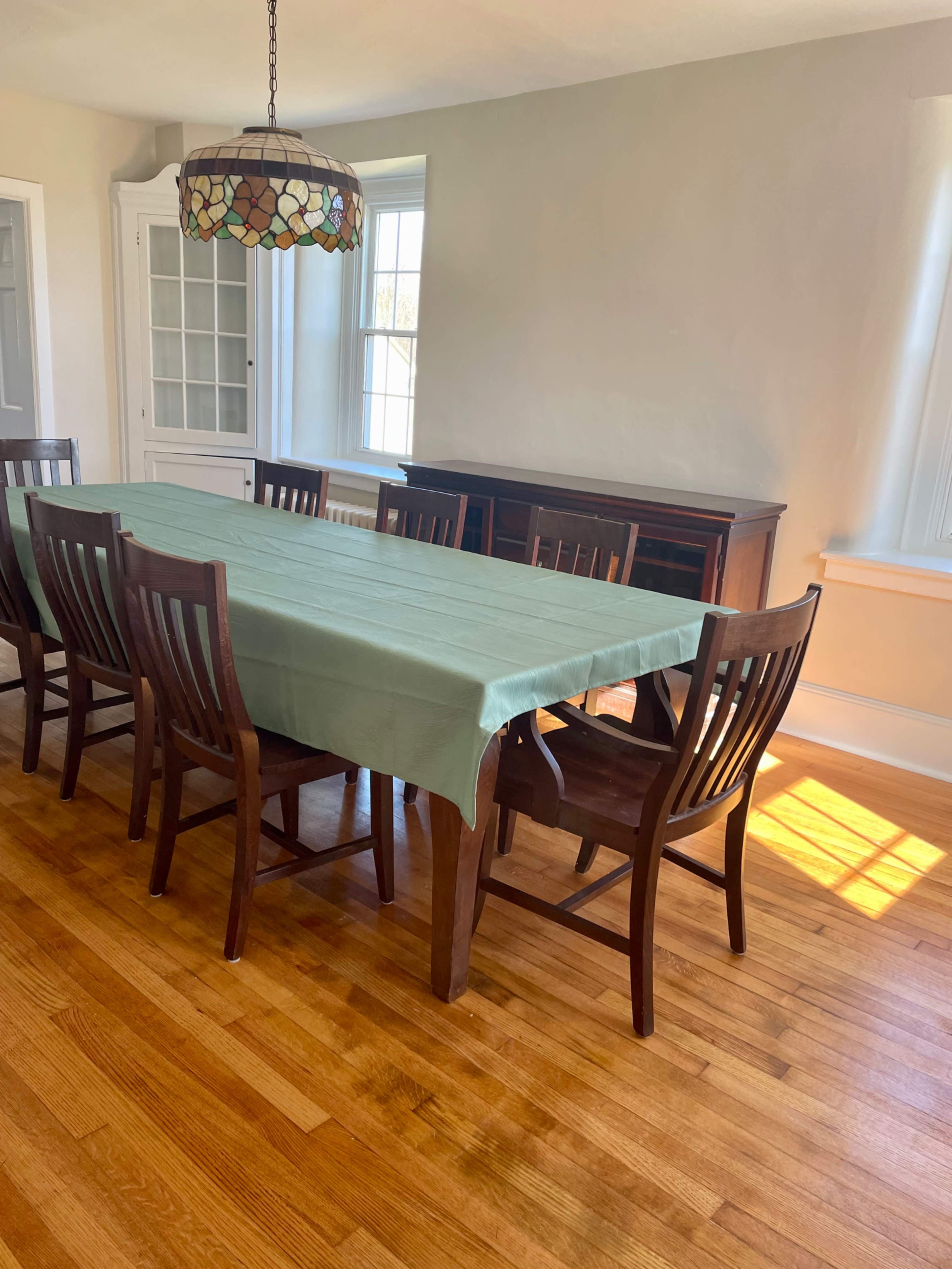 A long wooden dining table with a green tablecloth is surrounded by eight wooden chairs in a bright room with large windows.
