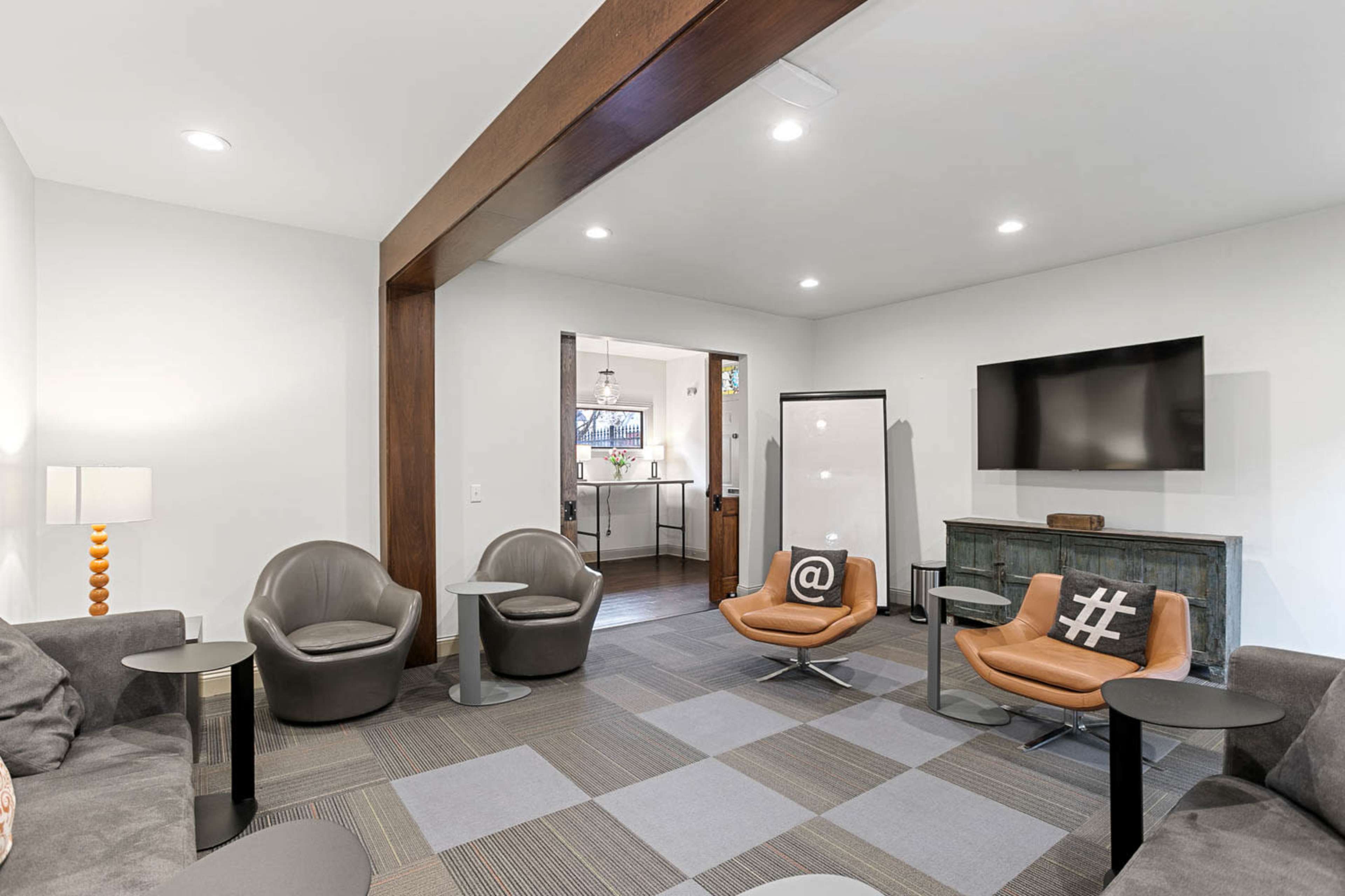 The image shows a modern lounge area with two stylish armchairs, a small table beside each, a sofa, and a large television mounted on the wall.