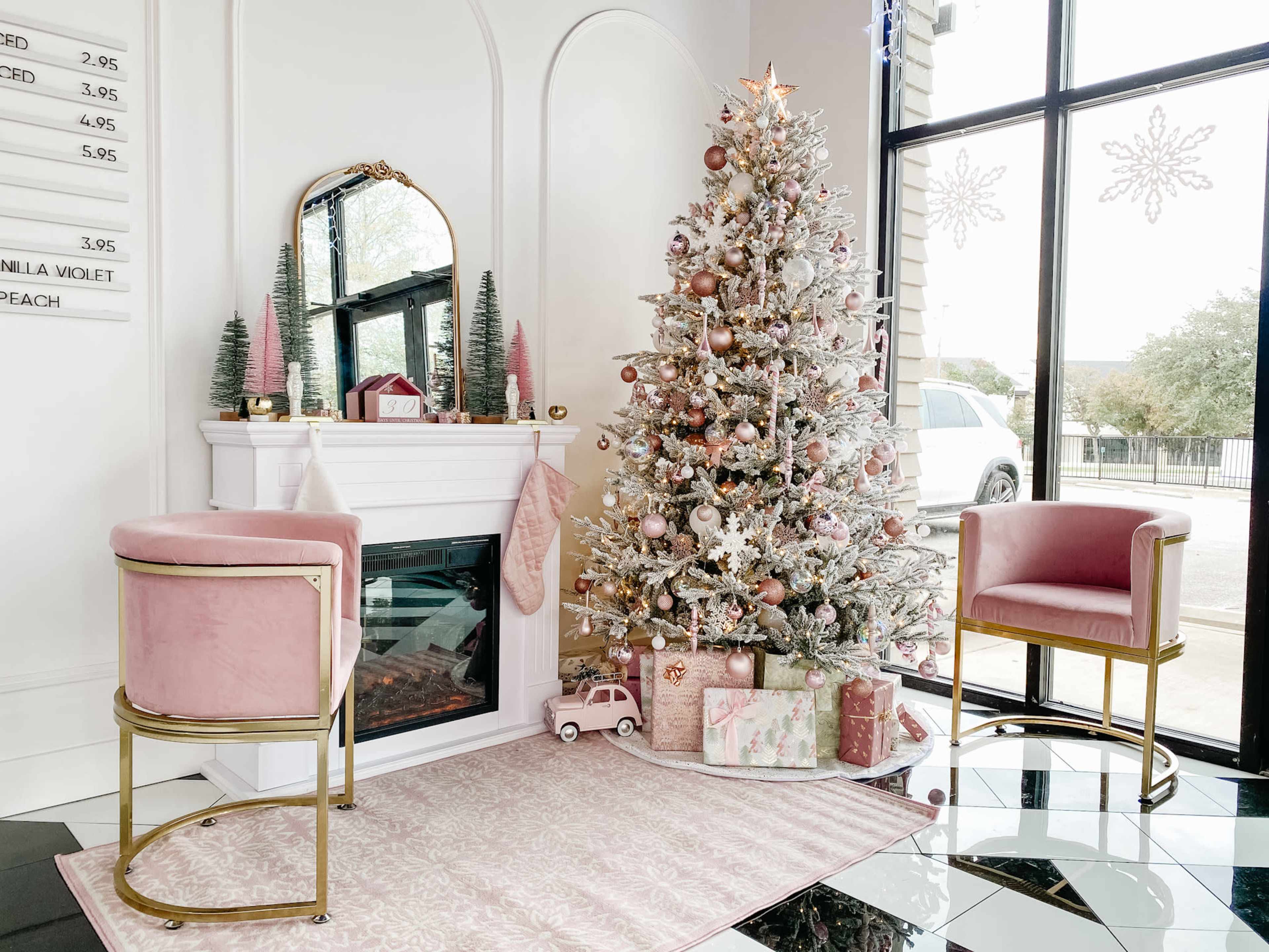 The scene features a decorated Christmas tree with pink ornaments beside a fireplace, flanked by two stylish chairs and a soft rug, all set in a bright room with large windows.