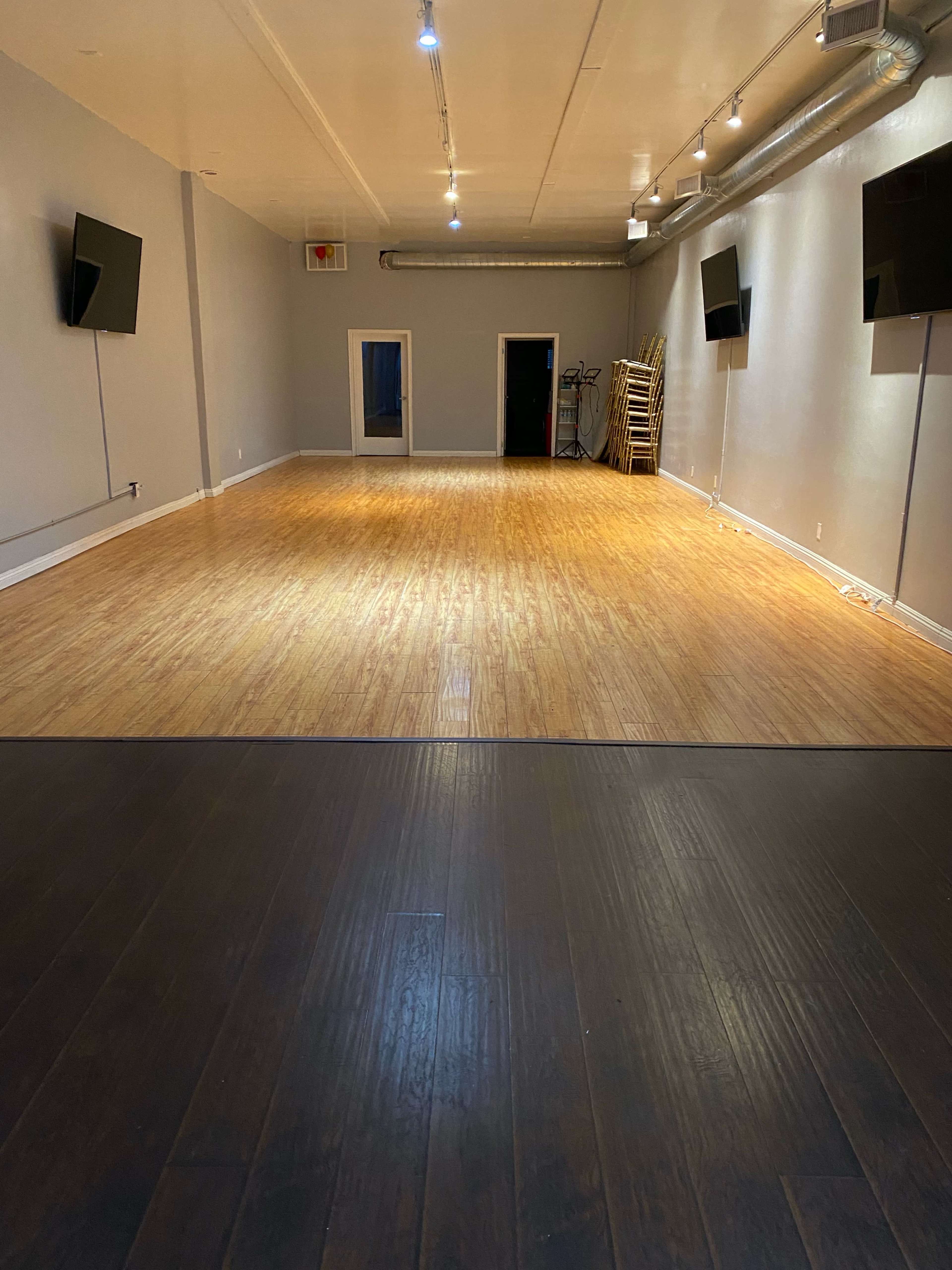 The image shows a spacious, well-lit room with wooden flooring and empty walls, featuring two mounted televisions and a door at the back.