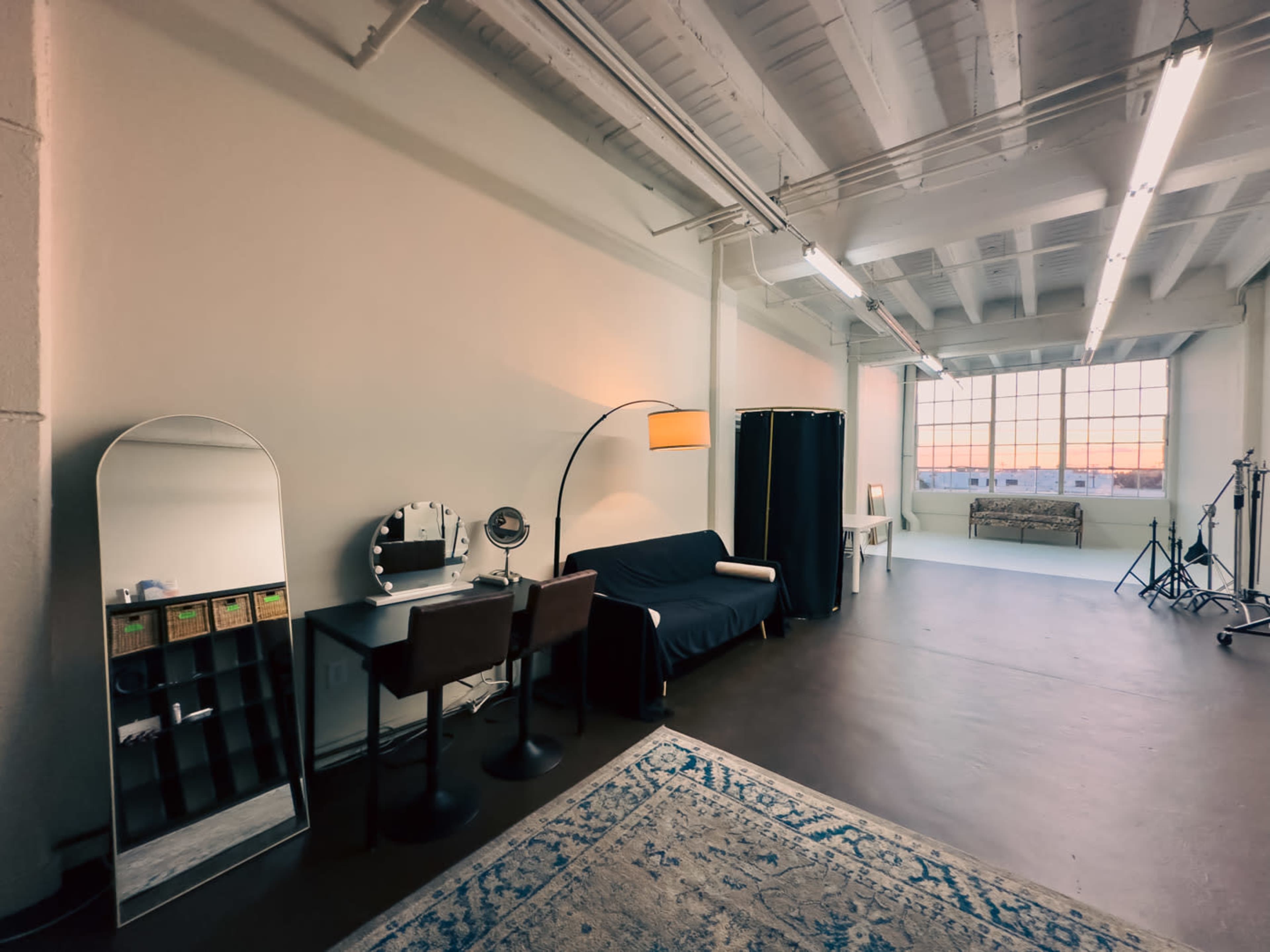 The image shows a spacious, minimalistic room featuring a dark sofa, a dressing table with a large mirror, and a window with sunset light illuminating the space.